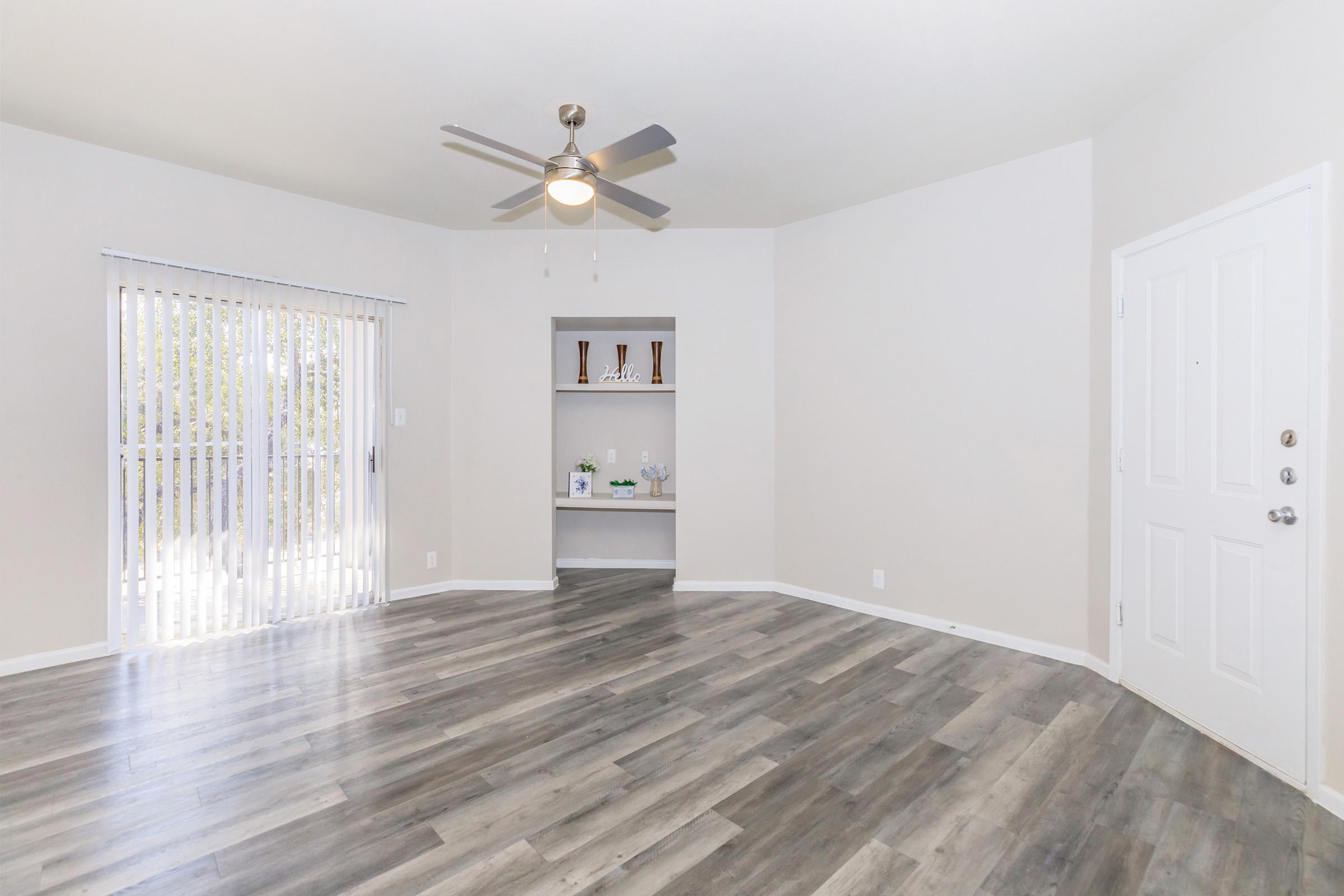 A bright and spacious living room with light gray walls and wood laminate flooring. There's a ceiling fan, a sliding glass door with vertical blinds, and a small nook with decorative shelves. The room is uncluttered and inviting, ideal for comfortable living.