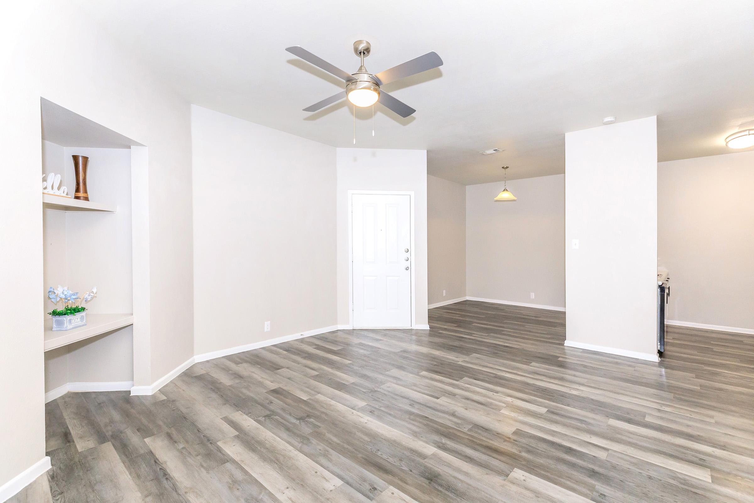 Spacious living room featuring gray wood-like flooring, white walls, and a ceiling fan. A shelving unit is on the left with decorative items. The front door is prominently positioned, with a light fixture above and a small pendant light in the background, creating a bright and inviting atmosphere.