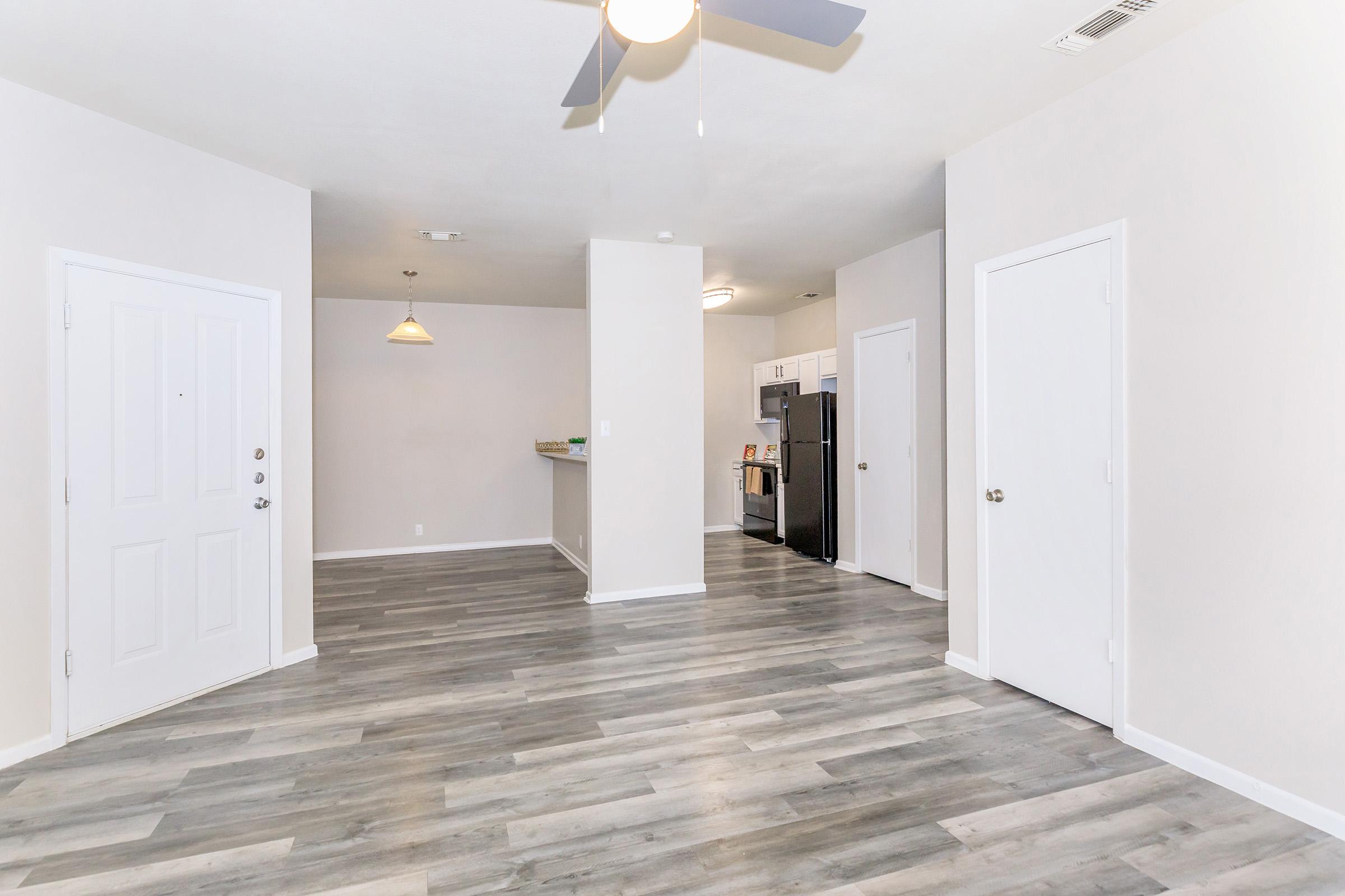 Spacious interior of a modern apartment featuring light gray walls and wood-like flooring. The open layout includes a living area with a ceiling fan, leading to a kitchen visible in the background. There are two white doors on the right and a door leading to the exterior on the left.