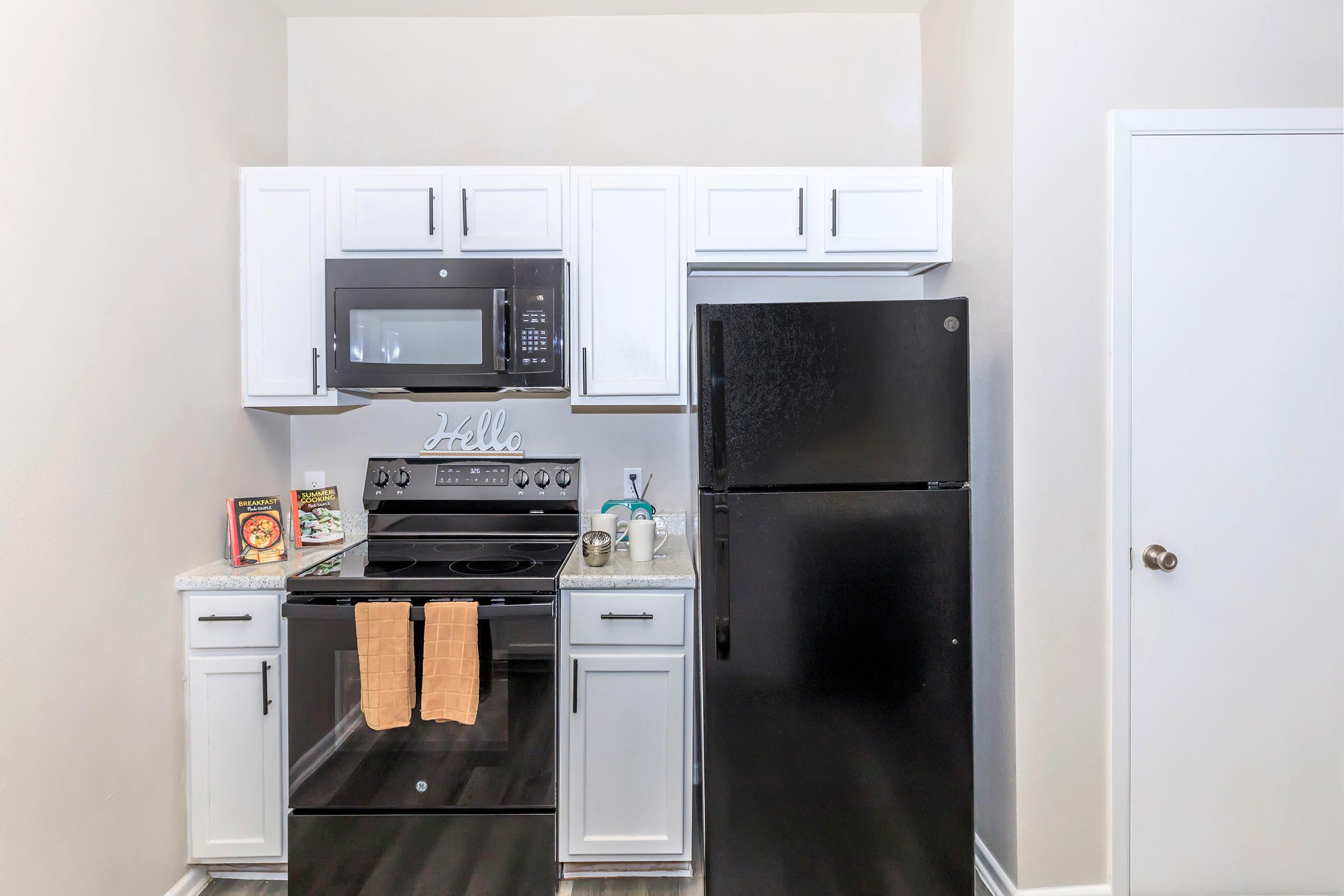 A modern kitchen featuring white cabinets, a black refrigerator, a stainless steel microwave, and an oven. Countertops have decorative items and cookbooks. Two tan towels hang from the oven. The walls are light-colored, and the flooring is dark, creating a stylish contrast.