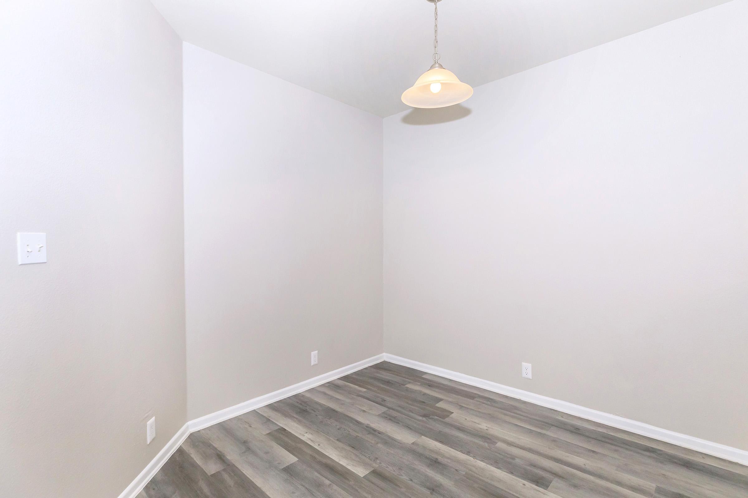 A small, empty room with light gray walls and a wooden floor. There is a single light fixture hanging from the ceiling, and no furniture or decorations are present. The space is well-lit, emphasizing the clean lines and neutral colors.