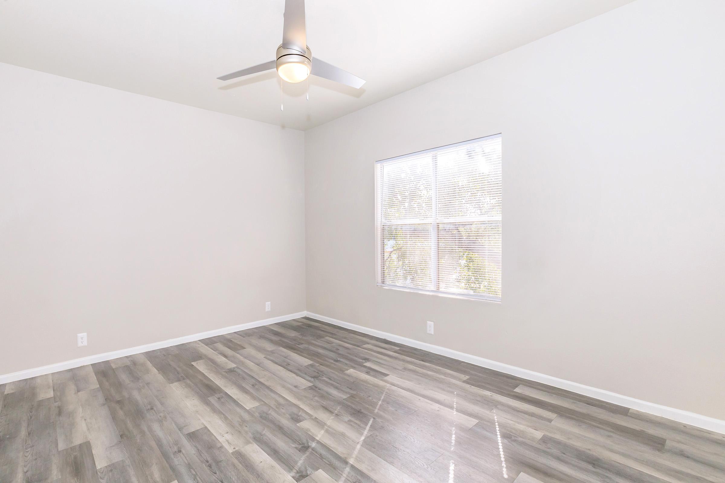 A bright, empty room with light gray walls and wood-patterned flooring. A ceiling fan is visible, and there is a window letting in natural light. The space is uncluttered, offering a minimalist and airy feel, making it suitable for various uses such as an office or bedroom.