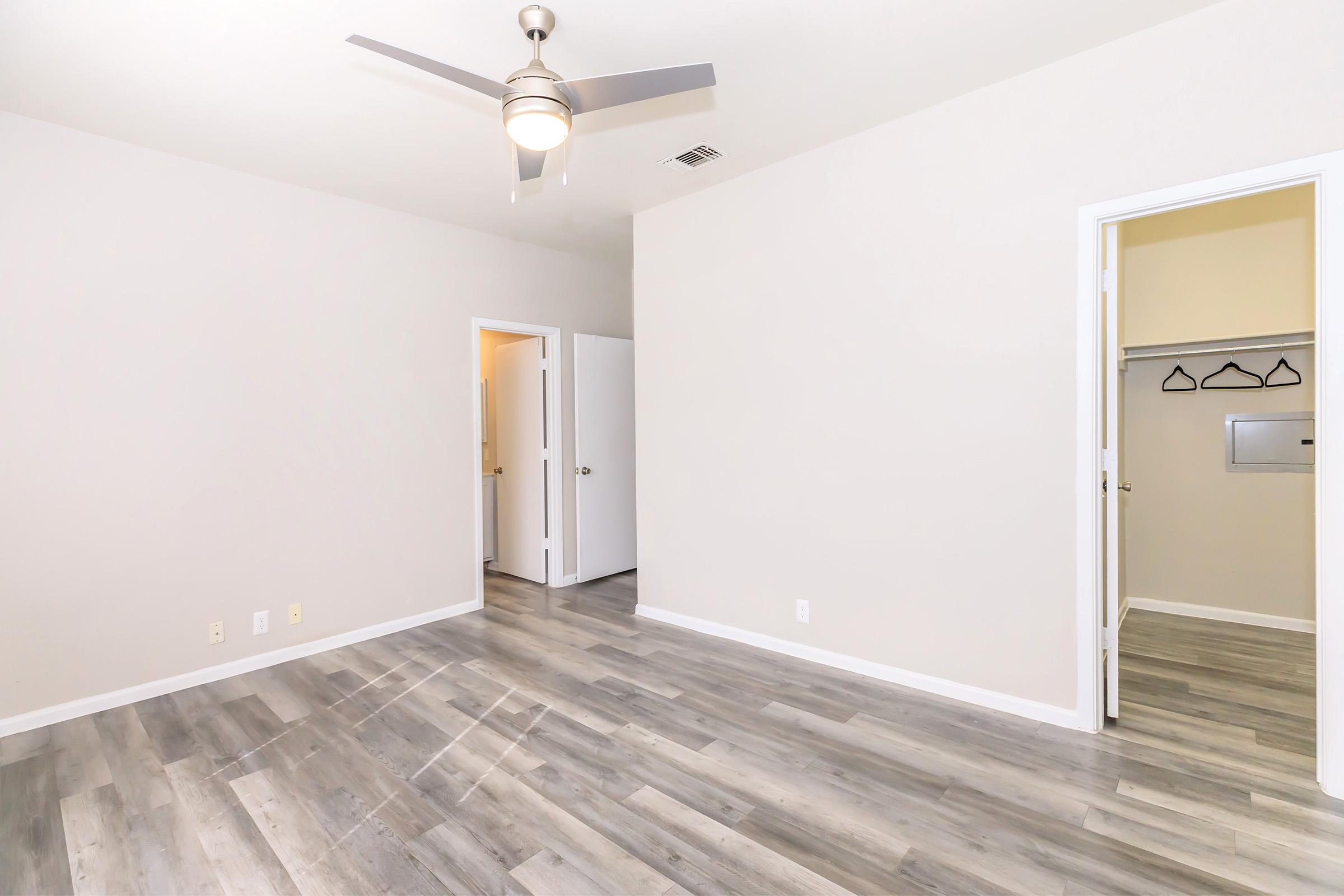 A spacious, empty room with light gray walls and light wooden flooring. There is a ceiling fan in the center, a doorway leading to another area, and a closet with hanging space visible. The overall atmosphere is bright and airy, ideal for personal or creative use.