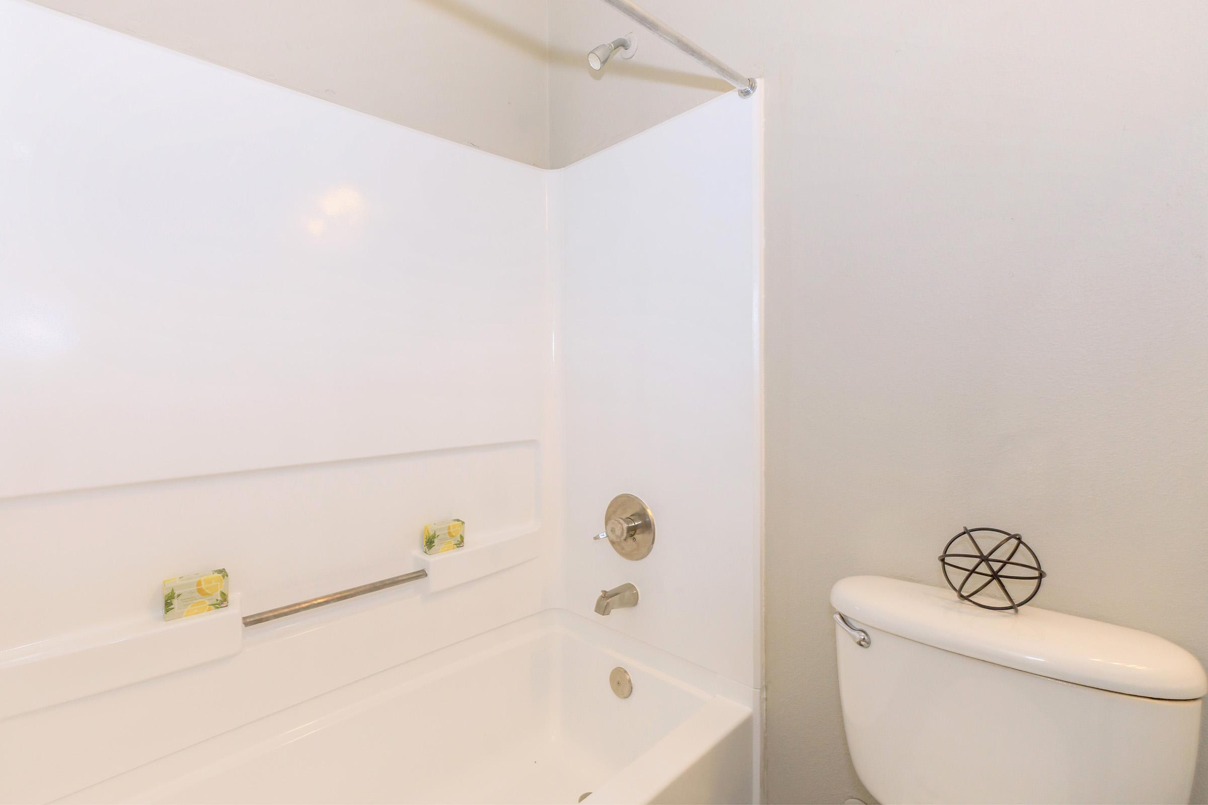 A clean and minimalistic bathroom featuring a white bathtub with a showerhead, a towel bar with two yellow towels, and a white toilet with an artistic black geometric decoration on top. The walls and fixtures are all white, creating a bright and airy atmosphere.