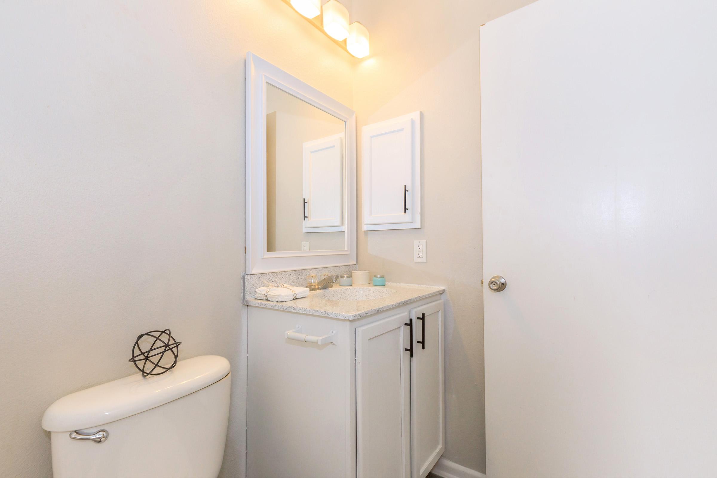 A well-lit bathroom featuring a white toilet, a clean countertop with a small decorative item, and a large mirror above a cabinet. The walls are painted in a soft gray, and there is a white door on the right side, contributing to a modern, minimalist aesthetic.