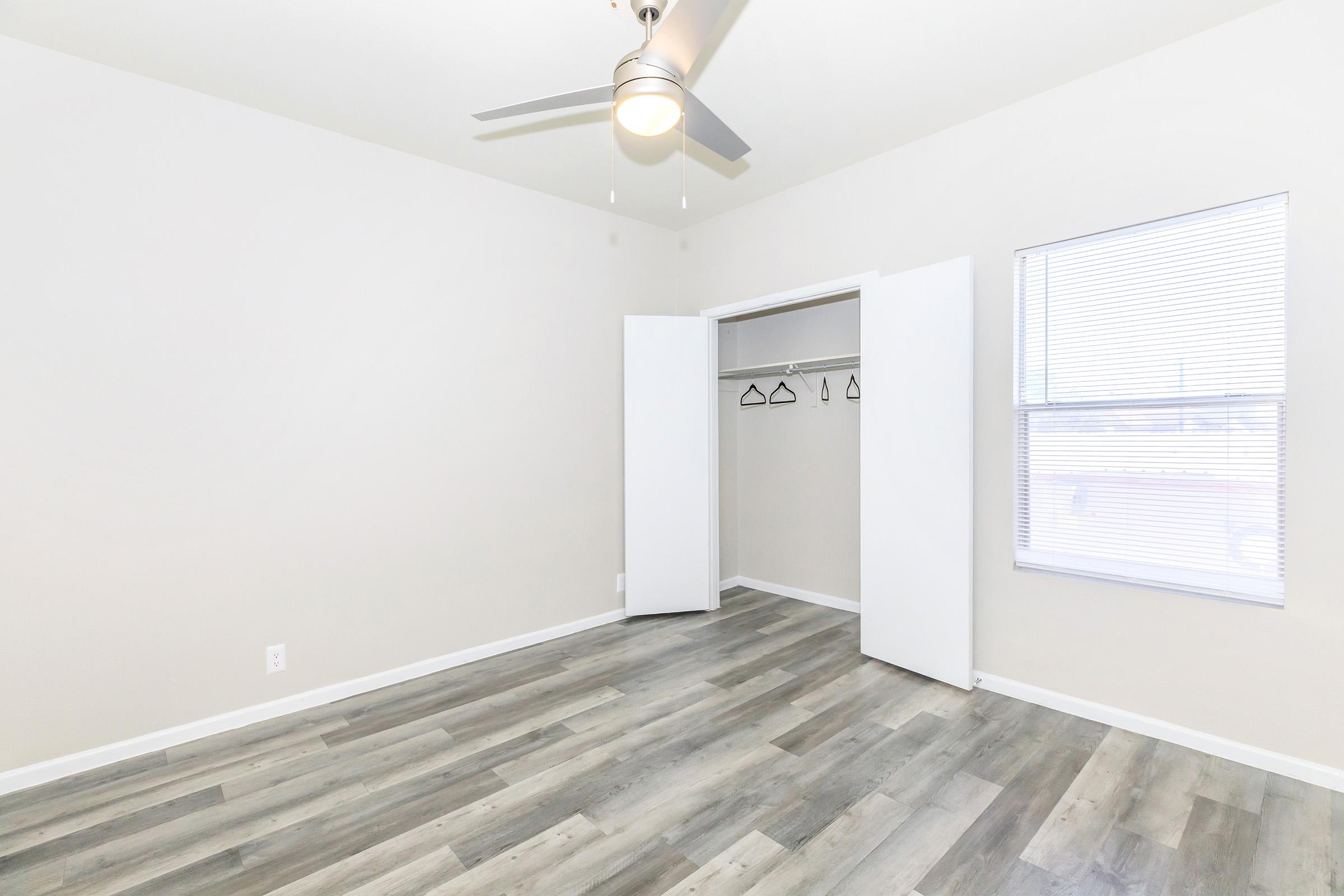 A spacious, empty room featuring light-colored walls and a ceiling fan. The floor is covered with gray wood-like planks. There is a closet with sliding doors on one side, and a window with blinds on the opposite wall, allowing natural light to enter.