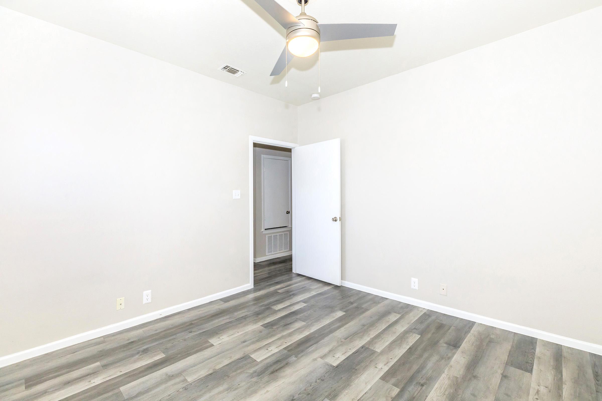 A minimalistic room featuring light-colored walls, a ceiling fan, and laminate flooring. A white door leads to another space, and there is no furniture present, giving the room an open and spacious feel. Natural light can be seen through the doorway.