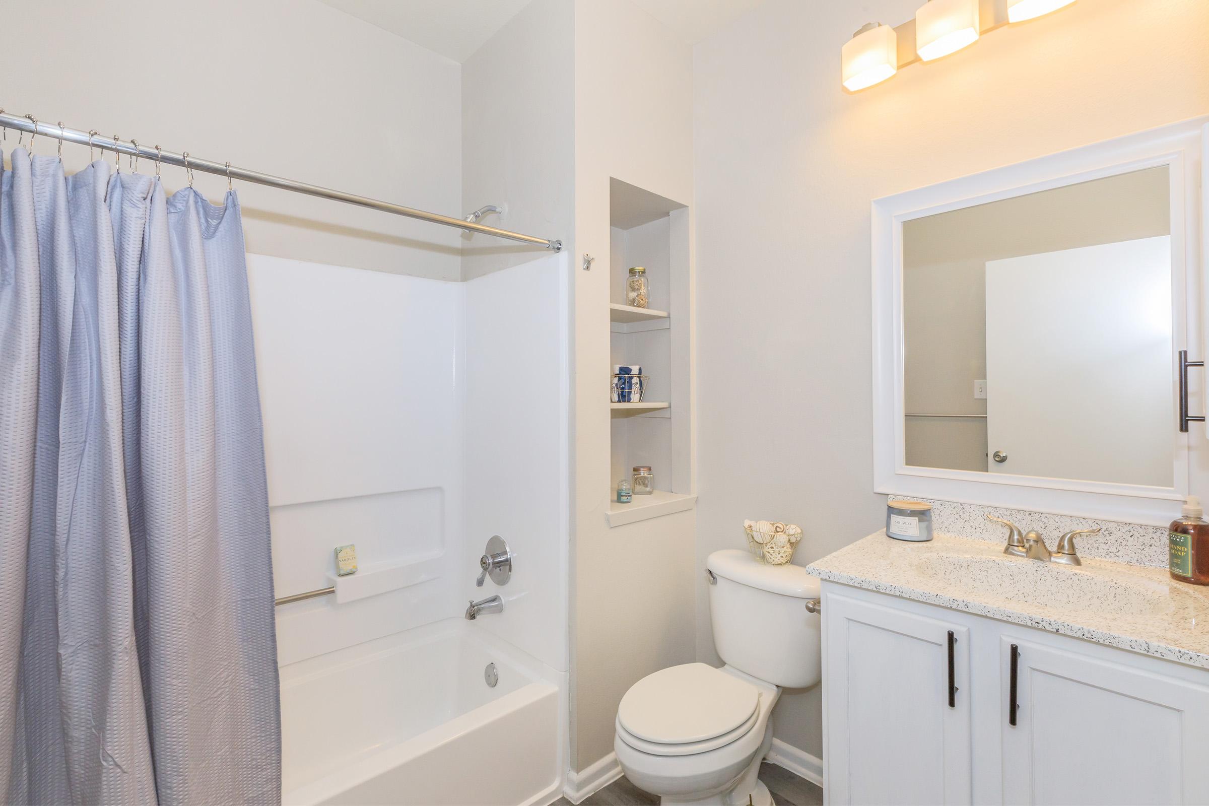 A modern bathroom featuring a bathtub with a shower curtain, a toilet, and a vanity with a granite countertop and a mirror. There are shelves built into the wall holding small decorative items and toiletries. Soft, neutral colors create a clean and inviting atmosphere.