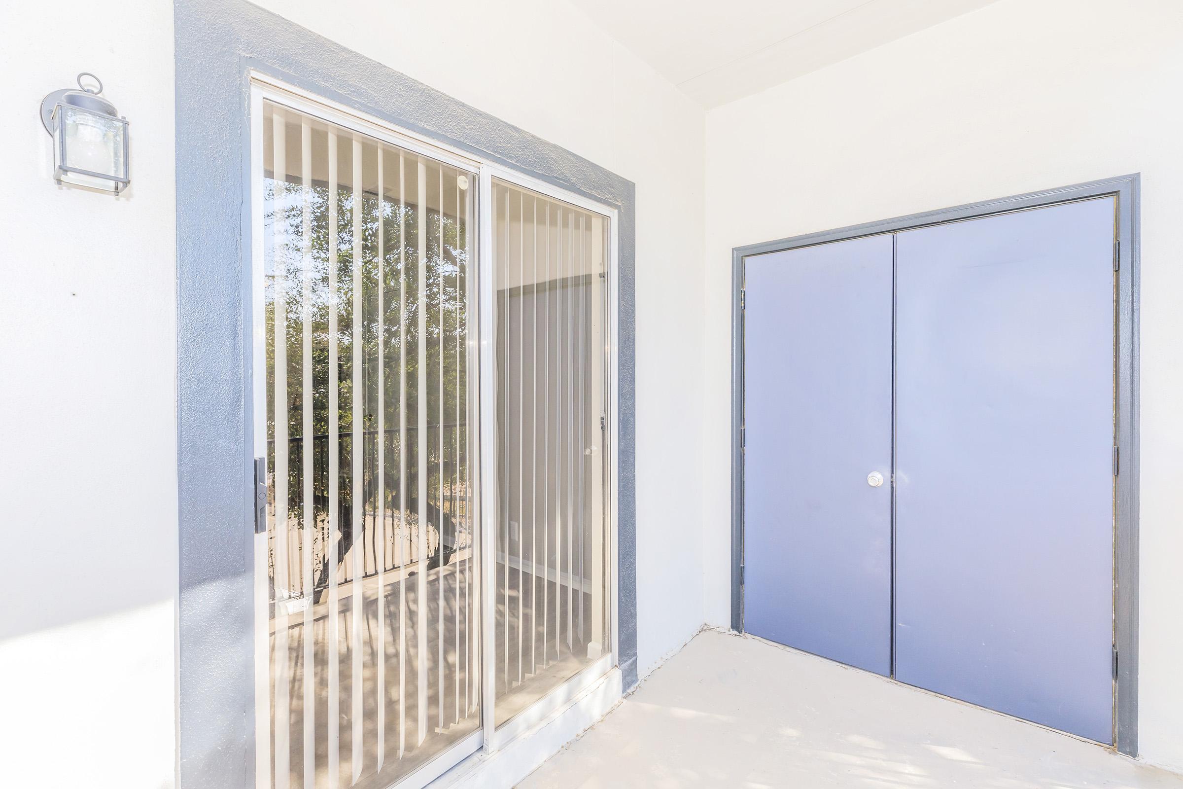 A well-lit entrance area featuring a glass sliding door with vertical blinds and a pair of blue double doors. The walls are painted white, and there's a wall-mounted light fixture beside the sliding door. The floor is unfinished or lightly textured, providing a clean, modern look.