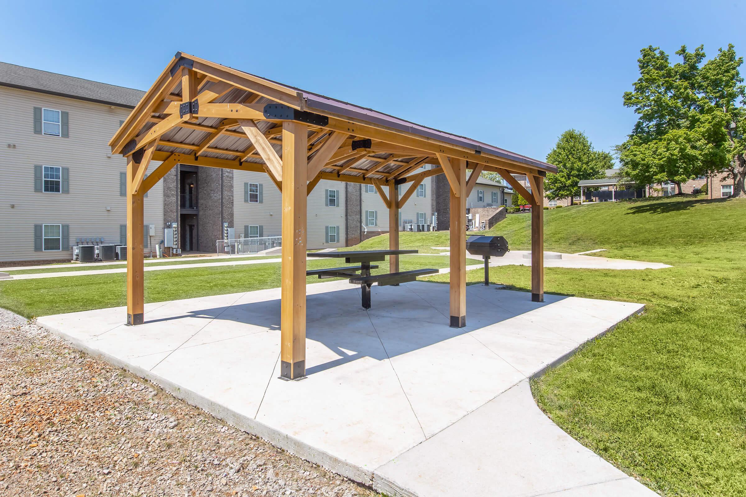 A wooden picnic shelter with a table and grill, surrounded by green grass and residential buildings in the background. The sky is clear and blue, and it appears to be a sunny day. The area is well-maintained, ideal for outdoor gatherings or relaxation.