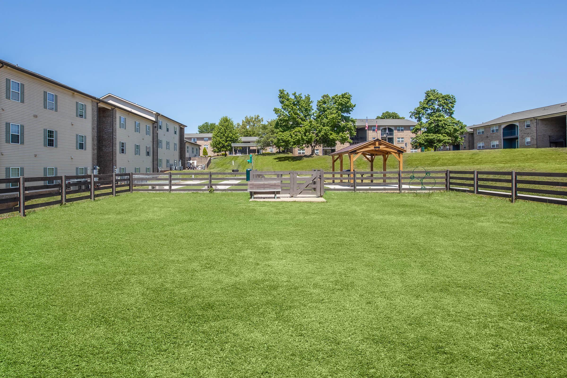 A spacious green courtyard with a well-maintained lawn, surrounded by wooden fencing. In the background, there are several multi-story apartment buildings. A shaded seating area is visible with a gazebo, creating an inviting space for relaxation and gatherings. The sky is clear and blue, enhancing the bright atmosphere.