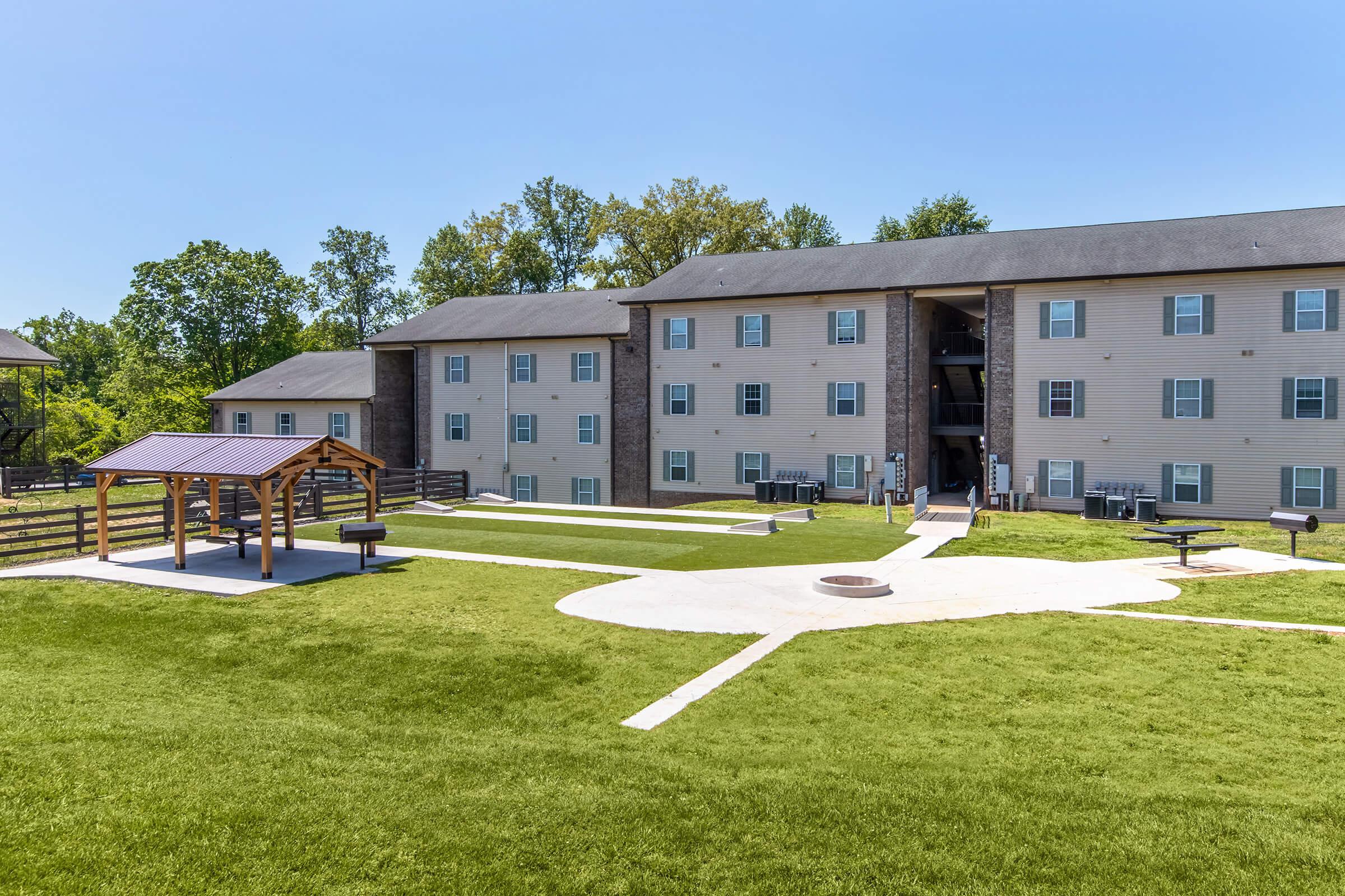 A well-maintained outdoor area featuring a grassy landscape, a picnic pavilion with benches, and a fire pit. In the background, a multi-story residential building with multiple units and nearby parking. Trees line the sides, providing a natural setting on a sunny day.