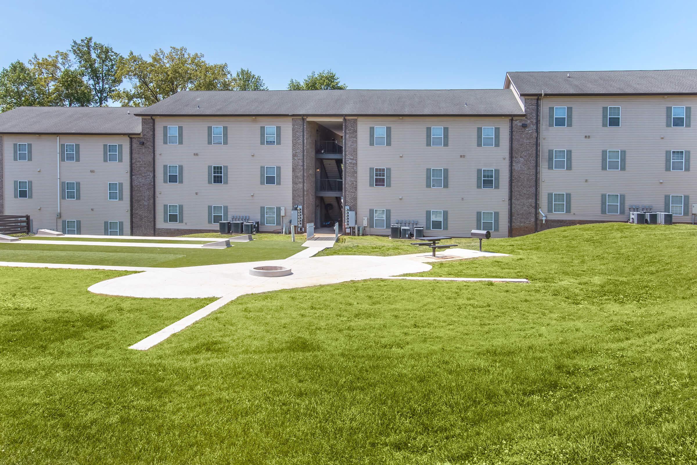 A multi-story residential building with light-colored siding and multiple windows. The structure is surrounded by well-kept green grass and a paved pathway leading to a communal area with seating. The sky is clear and blue, indicating a pleasant day.