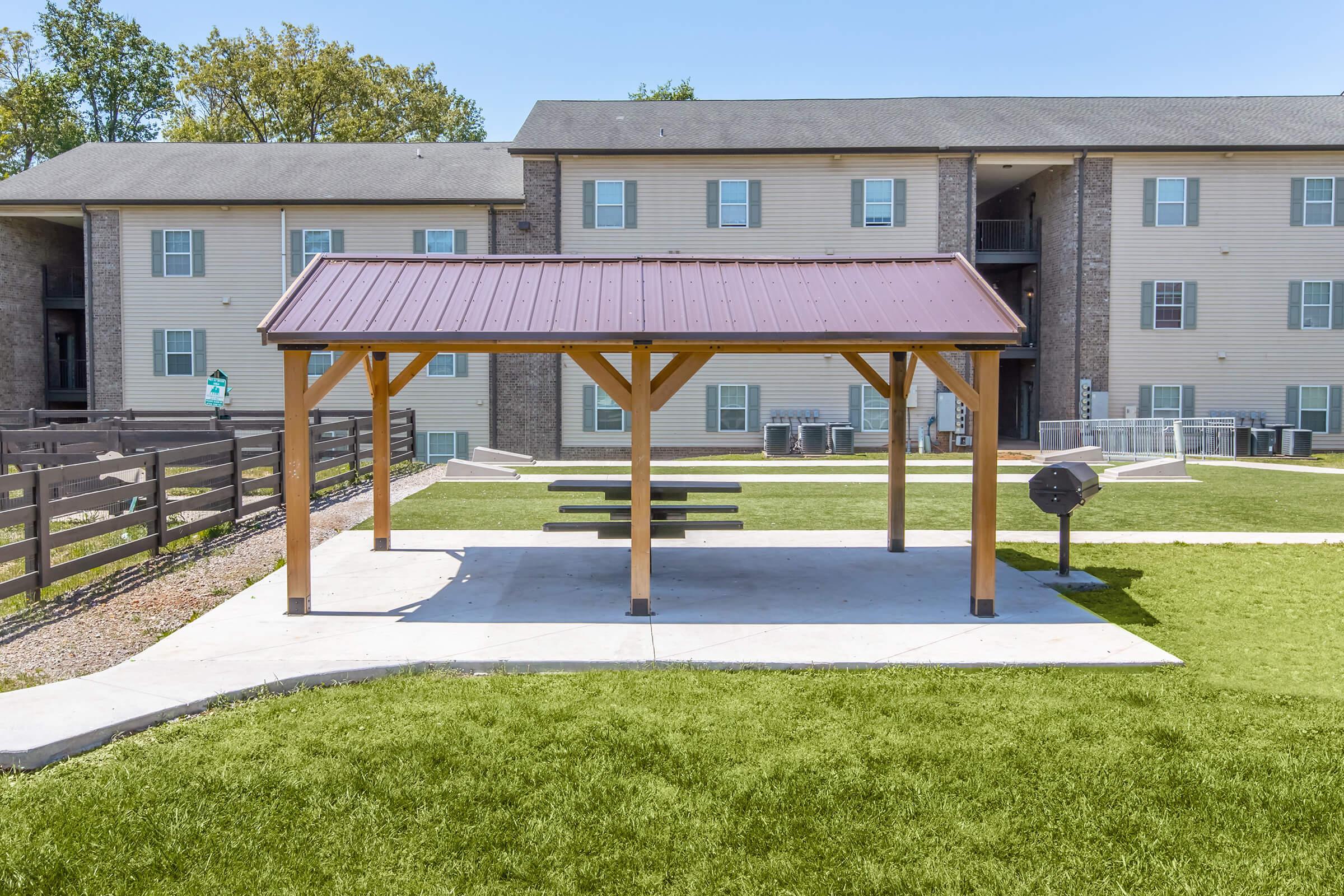 A wooden pavilion with a maroon roof is situated on a grassy area near an apartment building. Beneath the pavilion, there are picnic tables and a grill. In the background, multiple apartments can be seen, surrounded by well-maintained landscaping.