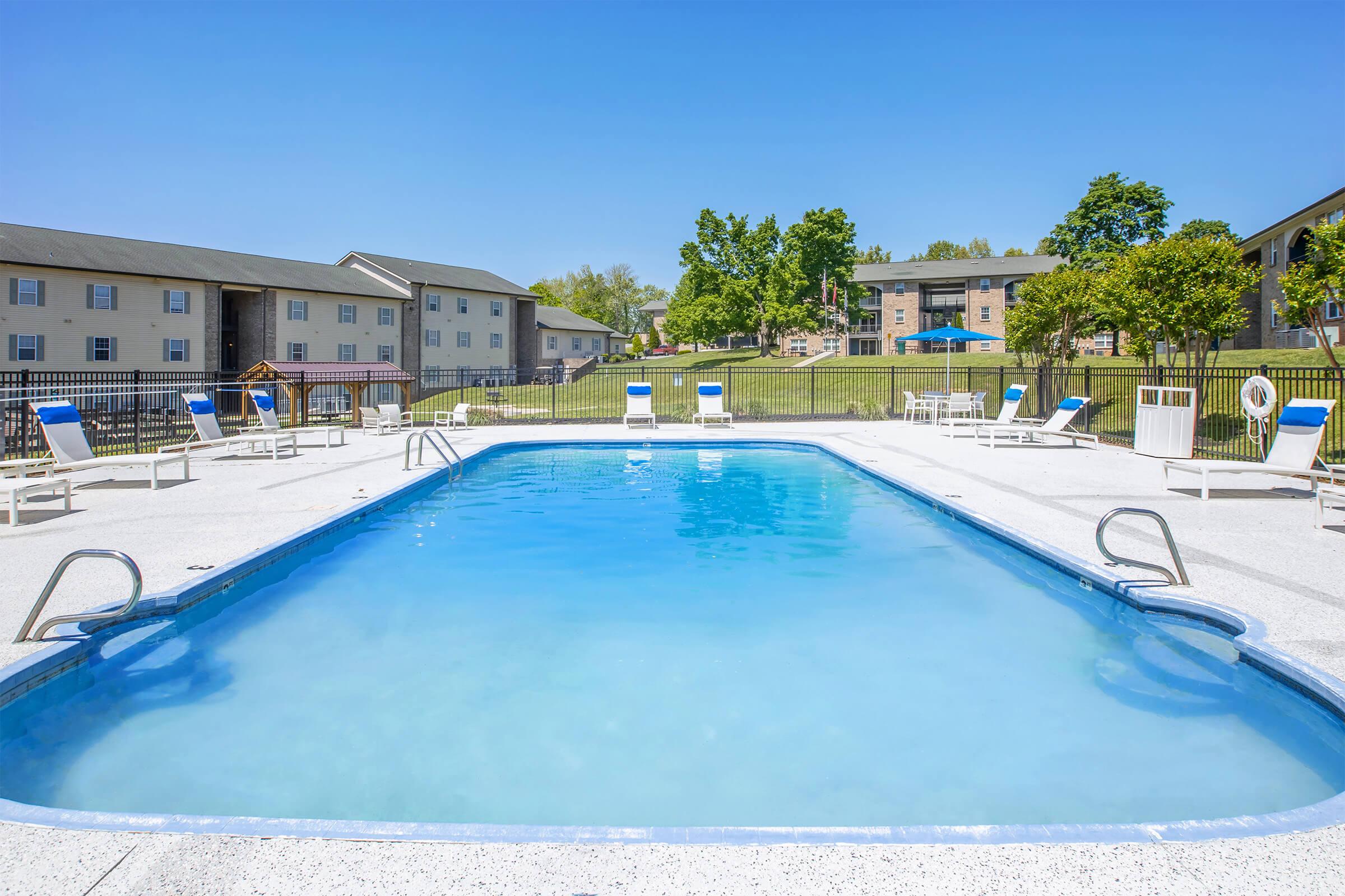 A clear swimming pool surrounded by lounge chairs and a fence, with two buildings and green trees in the background under a bright blue sky. The pool area is well maintained and inviting, perfect for relaxation and swimming.