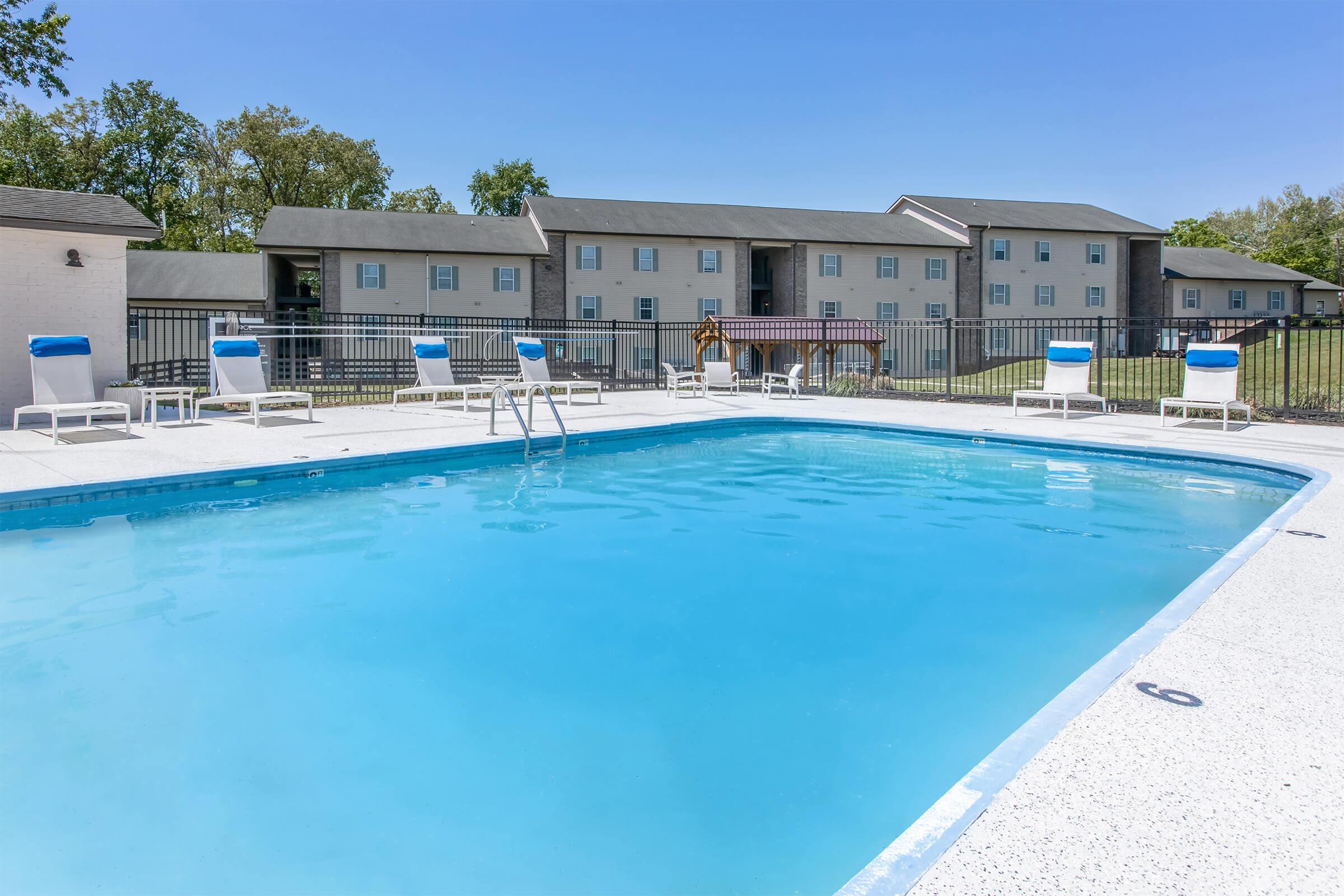 A bright blue swimming pool surrounded by lounge chairs, set in a well-maintained outdoor area. In the background, there are two apartment buildings with a grassy area, under a clear blue sky.