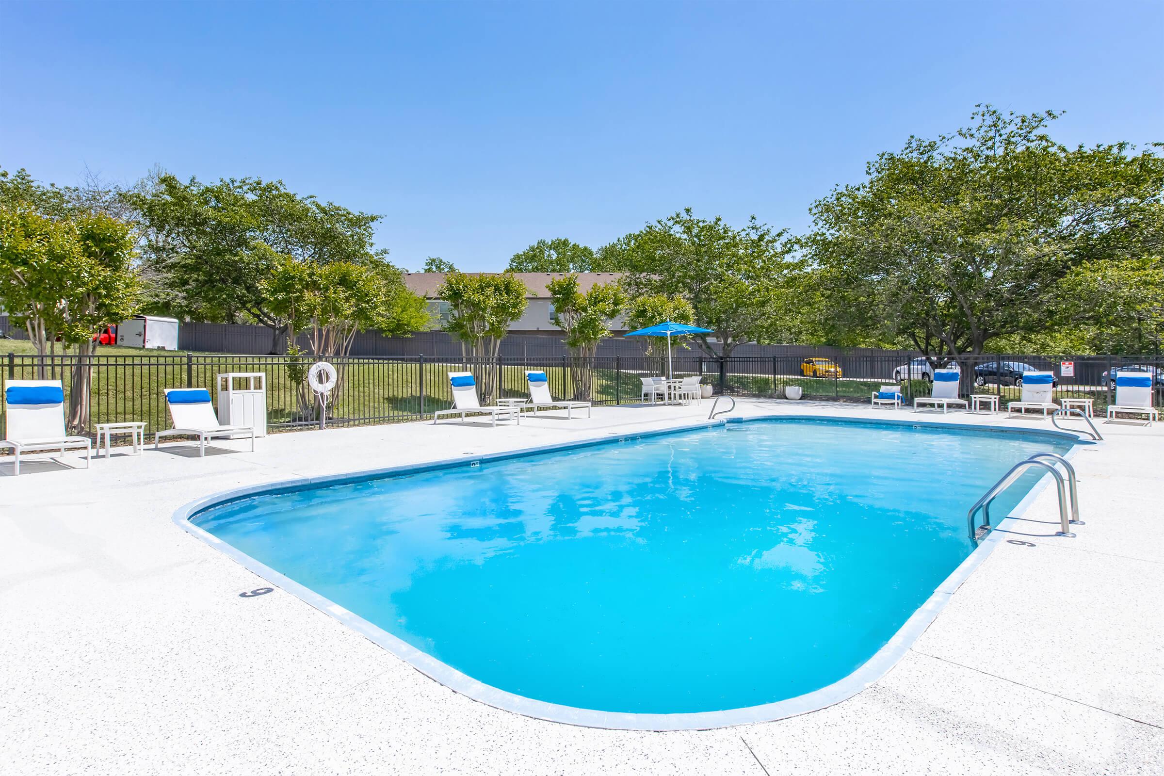 A clear blue swimming pool surrounded by lounge chairs. The area features trees in the background and a clear blue sky above, creating a relaxing outdoor atmosphere. A shaded umbrella stands near the pool, and there's a safety buoy on a fence nearby.