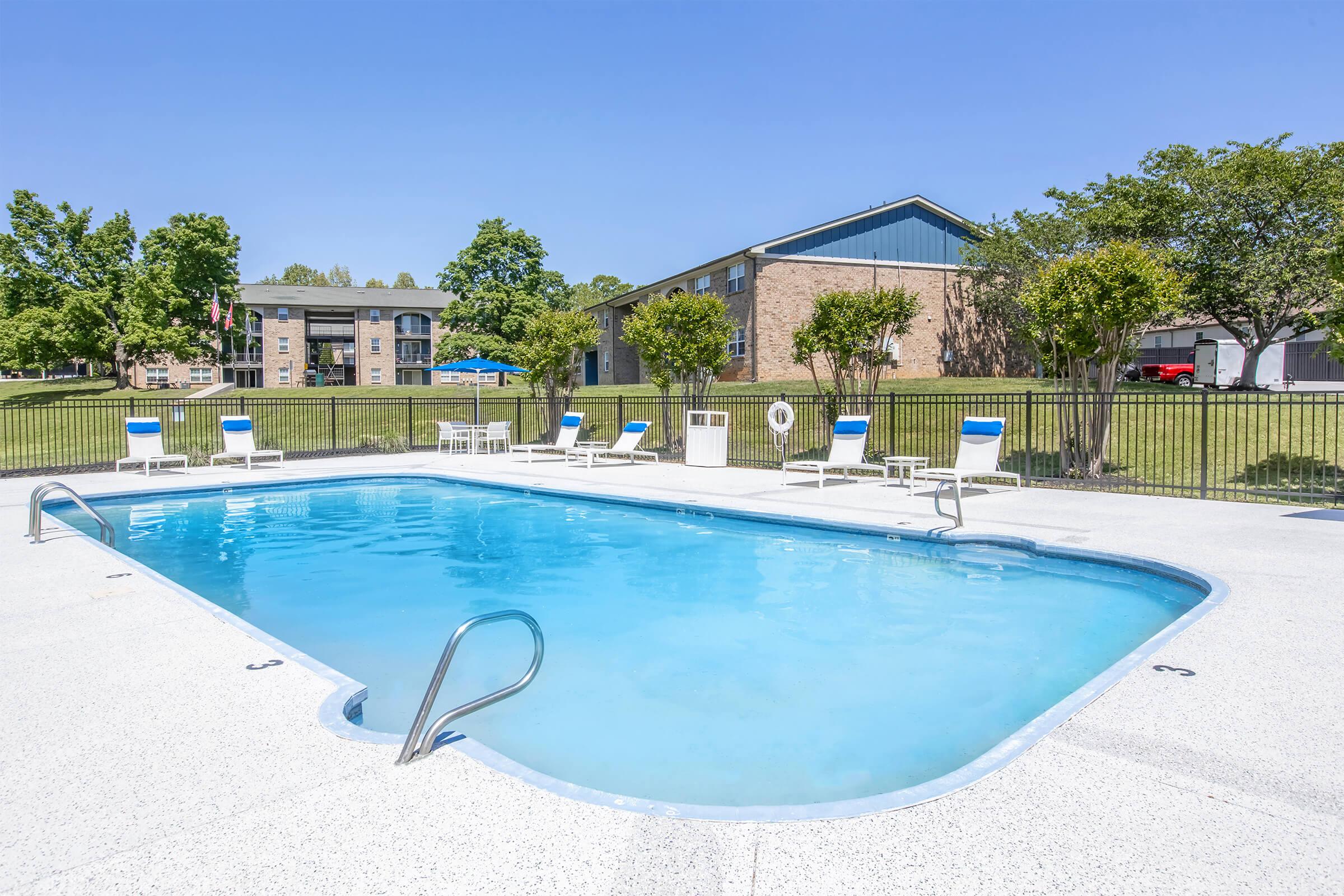A sunny outdoor swimming pool surrounded by lounge chairs. The pool has steps leading in, with a clean, blue water surface. In the background, there are trees and a building, contributing to a relaxing atmosphere perfect for leisure and recreation.