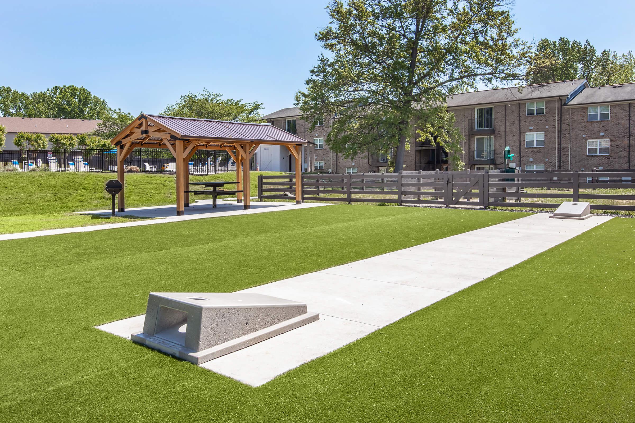 A well-maintained outdoor space featuring a synthetic grass area with a long, smooth surface for playing games. There is a shaded pavilion with picnic tables and a grill, and in the background, there are residential buildings with greenery. The scene is bright and inviting, perfect for social gatherings.