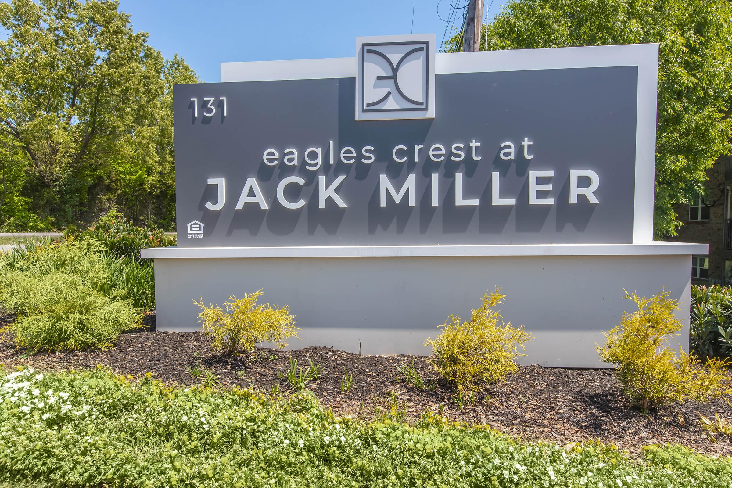 Sign for "Eagles Crest at Jack Miller" located at 131, featuring modern design elements and surrounded by landscaping with greenery and flowers. The sign is prominently displayed against a clear blue sky.