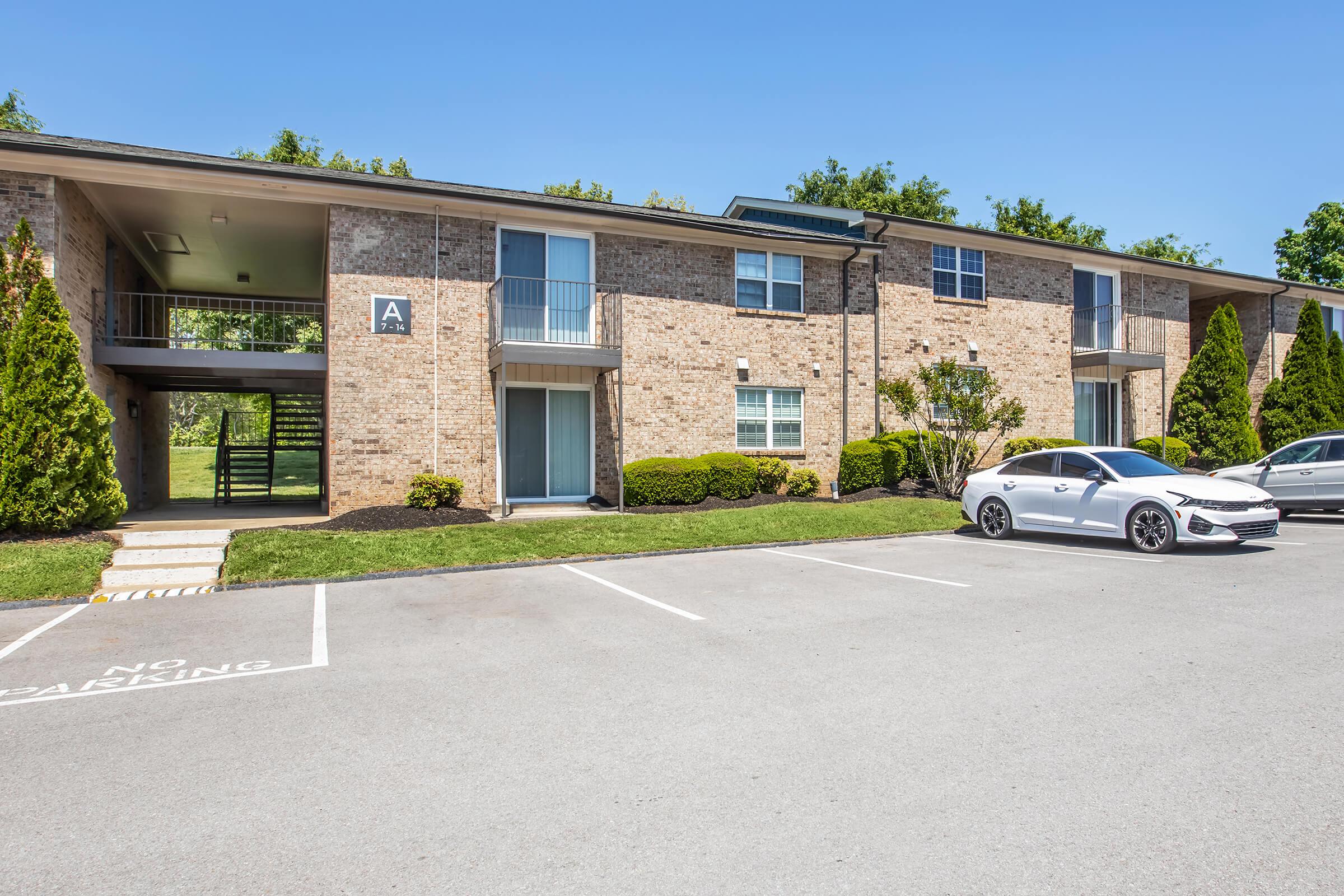 Exterior view of a brick apartment building with two floors, featuring a set of stairs leading to the upper level. The parking lot in front has several parking spaces, one of which is occupied by a white car. Lush green bushes and trees are visible around the building, set against a clear blue sky.