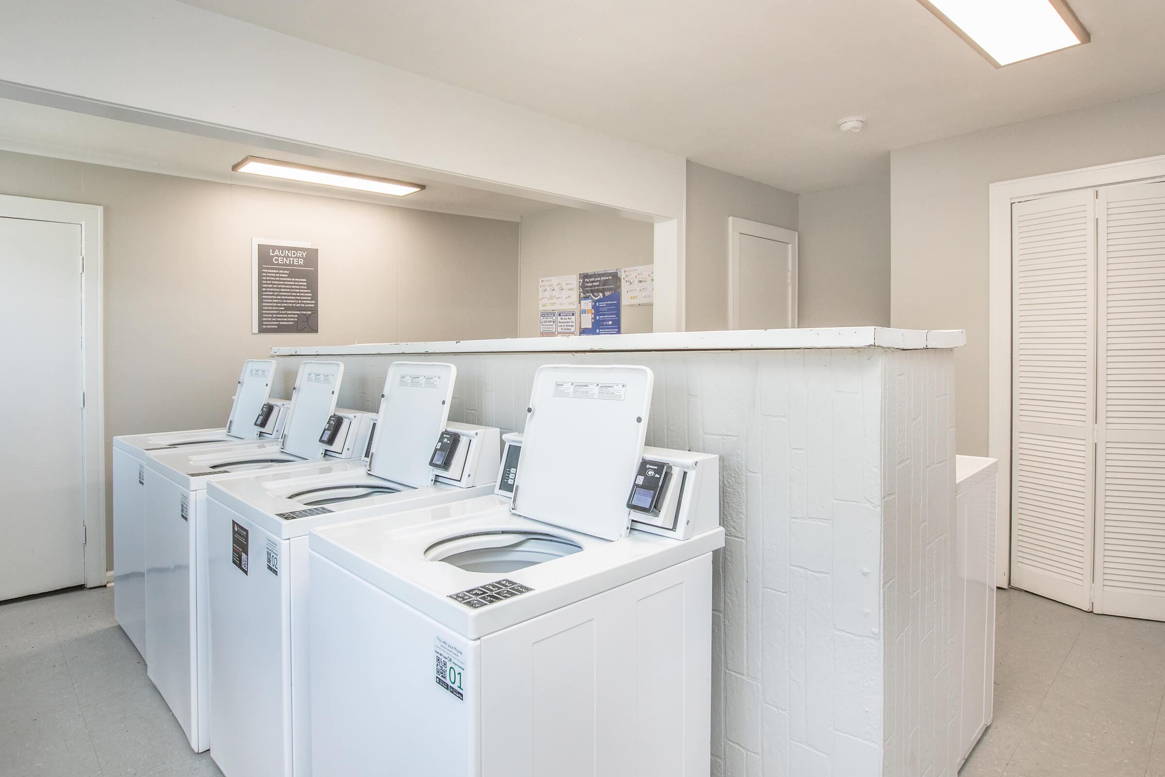 A bright, modern laundry room featuring several white washing machines lined up against a light gray wall. There is a divider separating the machines from a seating area, and informational signs are displayed on the wall. The floor is light gray, and the overall atmosphere is clean and organized.