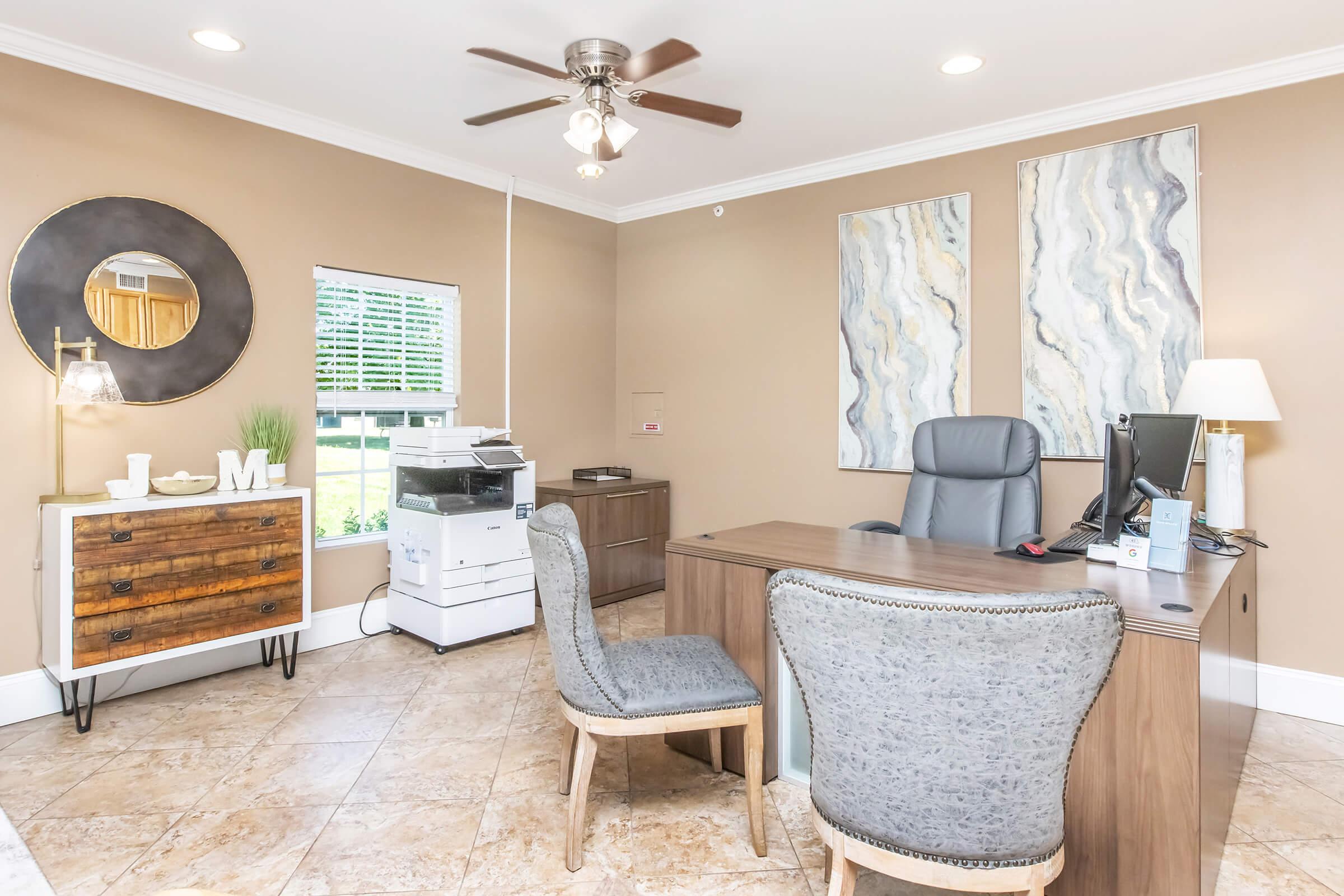 A well-lit office space featuring a wooden desk with a chair, a modern printer, and decorative wall art. A round mirror hangs on one wall, while a small table displays decorative items. The room has beige walls and tiled flooring, creating a welcoming atmosphere.