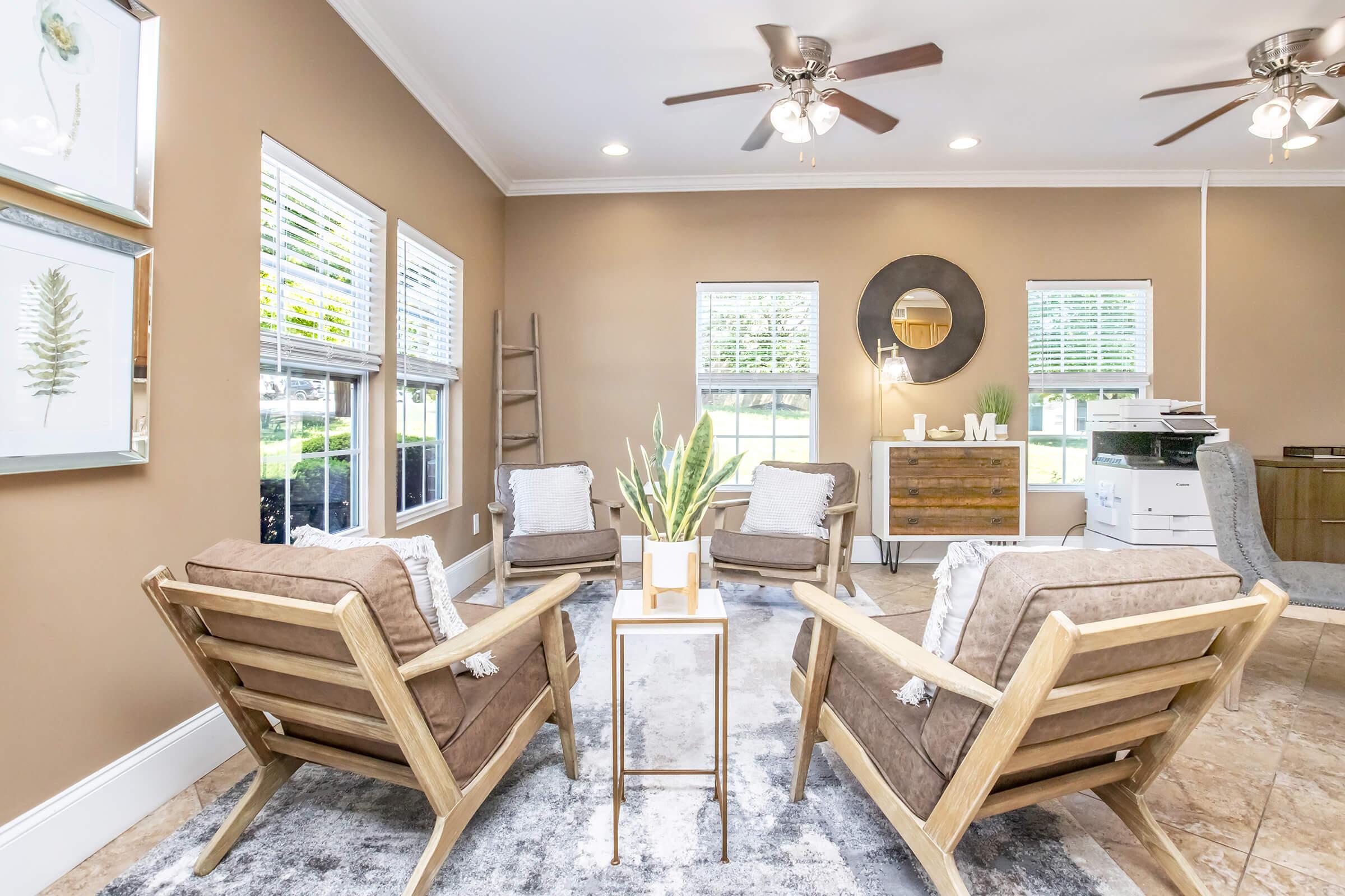 A cozy living room featuring two wooden armchairs with cushions, a small white table with a decorative plant, and a modern circular mirror on the wall. The room has large windows allowing natural light, a soft area rug, and a subtle earthy color scheme, creating a warm and inviting atmosphere.