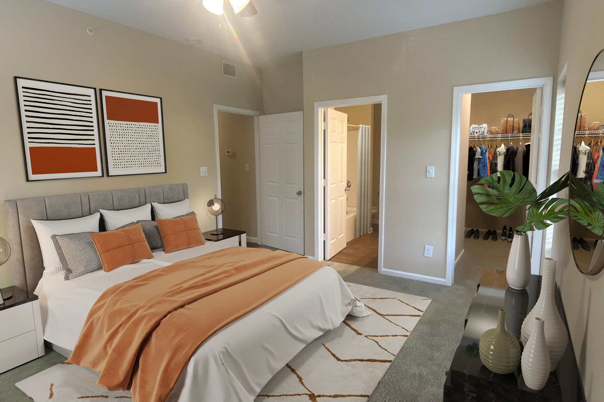 A cozy bedroom featuring a large bed with orange and gray bedding, decorative pillows, two framed art pieces on the wall, and a bedside lamp. There is a green plant by the bed, light-colored walls, and a carpeted floor. A doorway leads to a walk-in closet, and another door opens to a bathroom.