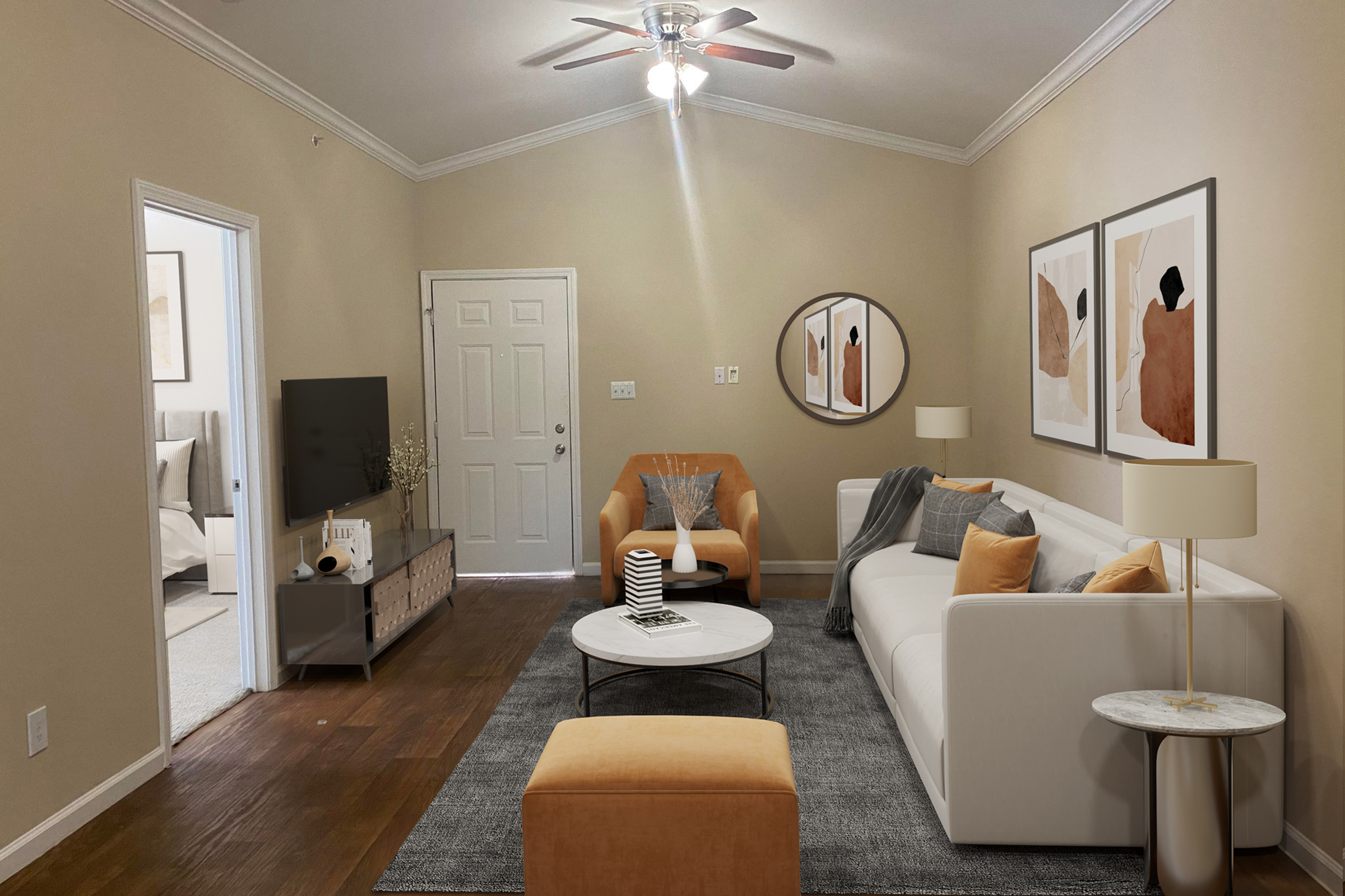 Cozy living room featuring a light-colored sofa with decorative pillows, a round coffee table, and an orange accent chair. A TV stand with a television is positioned across the room. The walls are painted in a warm tone, complemented by framed abstract art and a circular mirror. A doorway leads to a bedroom area.