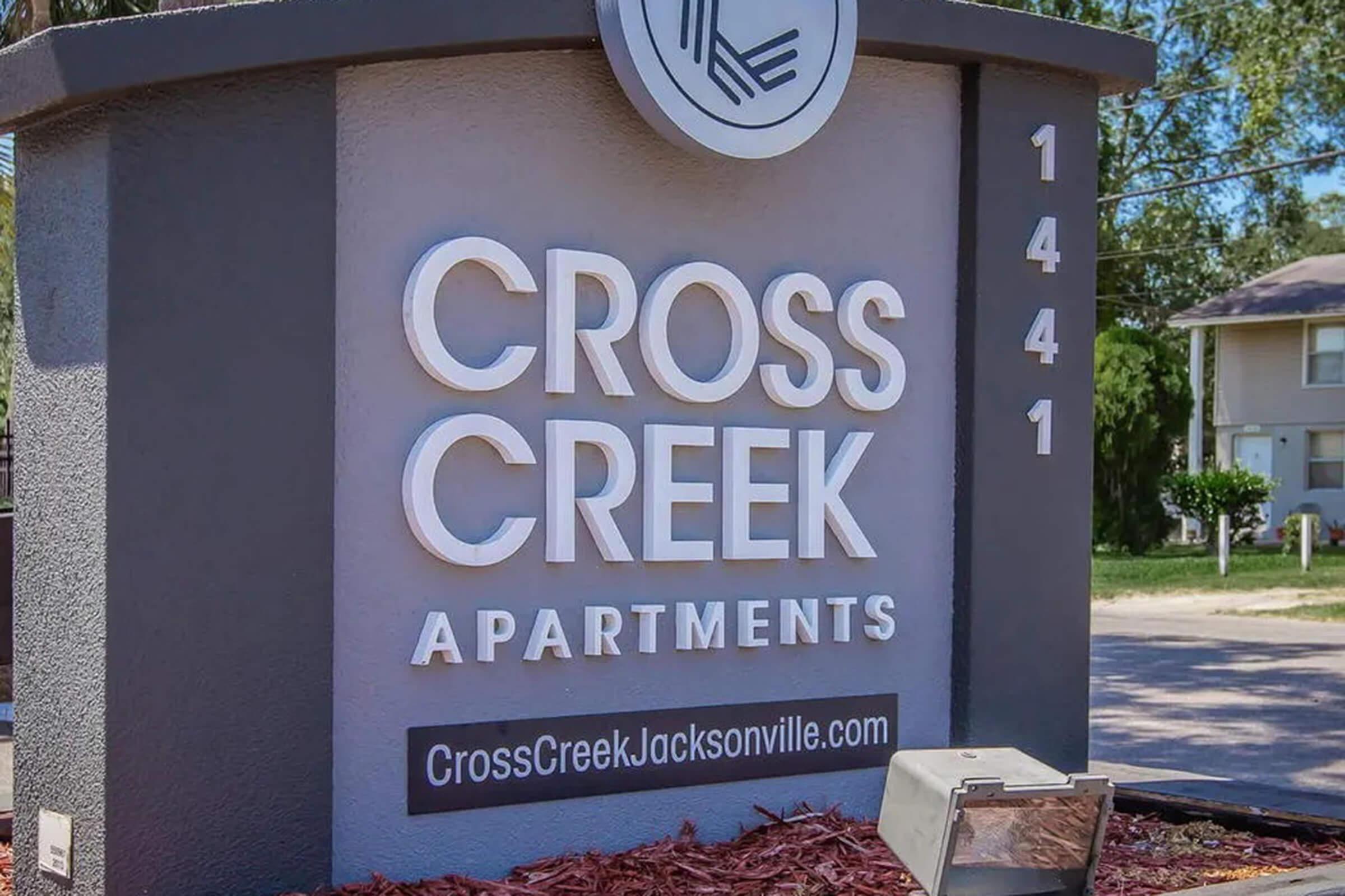 Sign for Cross Creek Apartments located at 1441, featuring the name in large, modern lettering. The background is a dark color with a minimalist design. A small box at the base of the sign appears to house lighting or a mailbox, surrounded by mulch.
