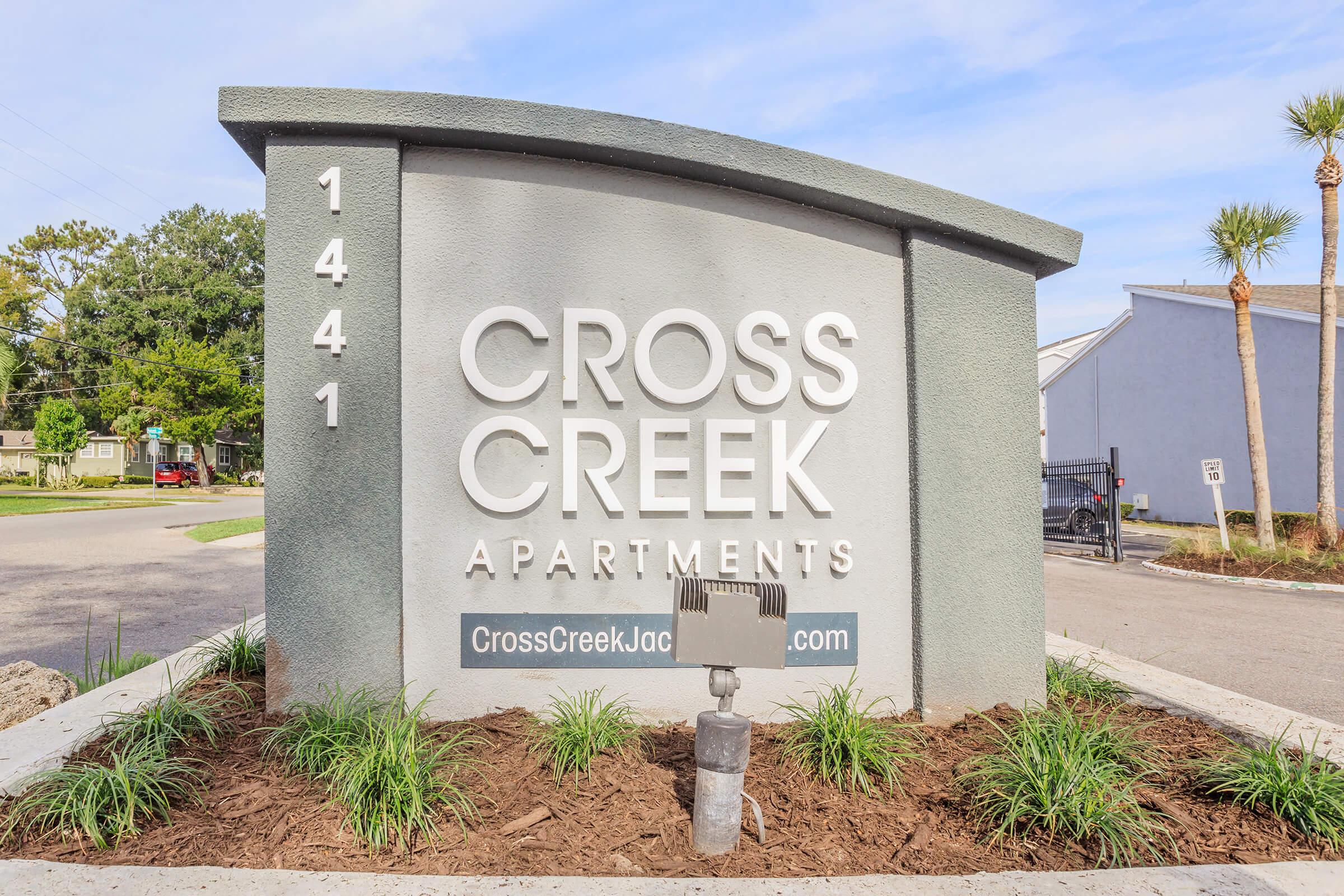 Sign for Cross Creek Apartments located at 1441, featuring the name "CROSS CREEK APARTMENTS" prominently displayed. The sign is modern and welcoming, surrounded by green landscaping and palm trees, with a website link at the bottom. Clear blue sky in the background.
