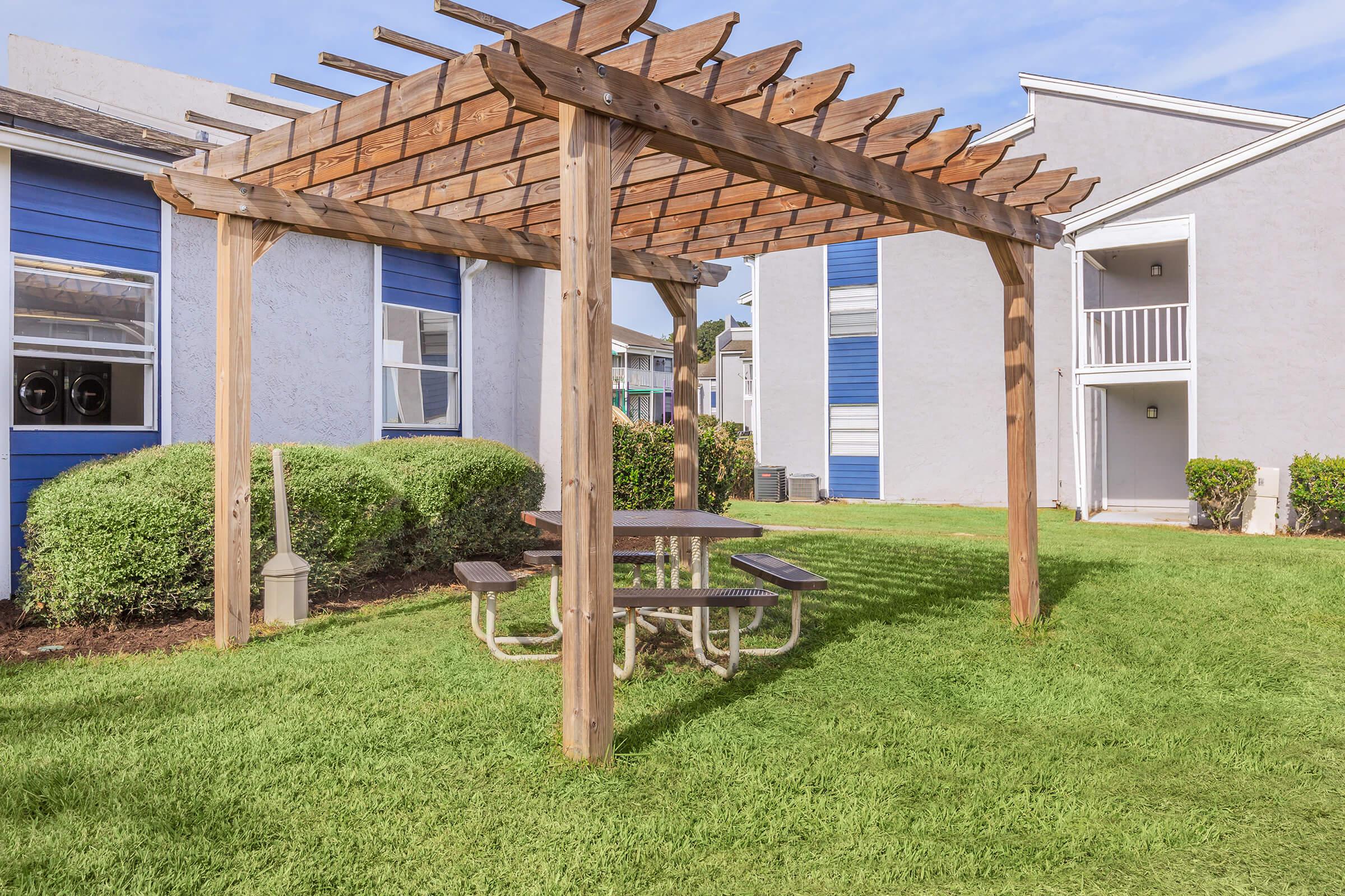 A wooden pergola with a picnic table underneath, surrounded by green grass and well-kept shrubs. In the background, there are two blue and gray apartment buildings, providing a cozy outdoor space for relaxation.