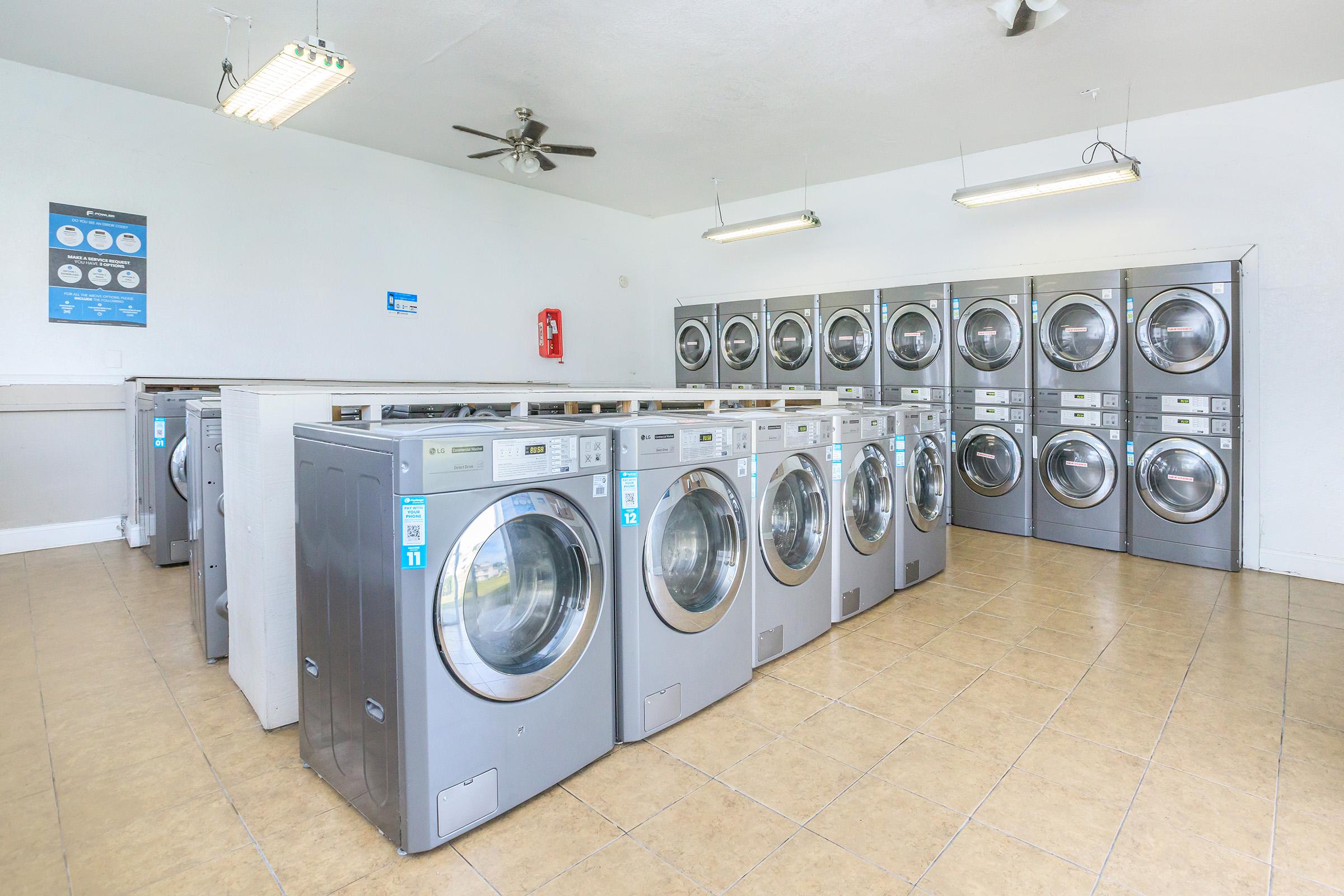 A clean and well-lit laundromat featuring multiple front-loading washing machines and dryers lined up against the wall. The floor is tiled, and there are overhead lights and a ceiling fan. A poster with laundry instructions is visible on the wall, contributing to the tidy and organized atmosphere.