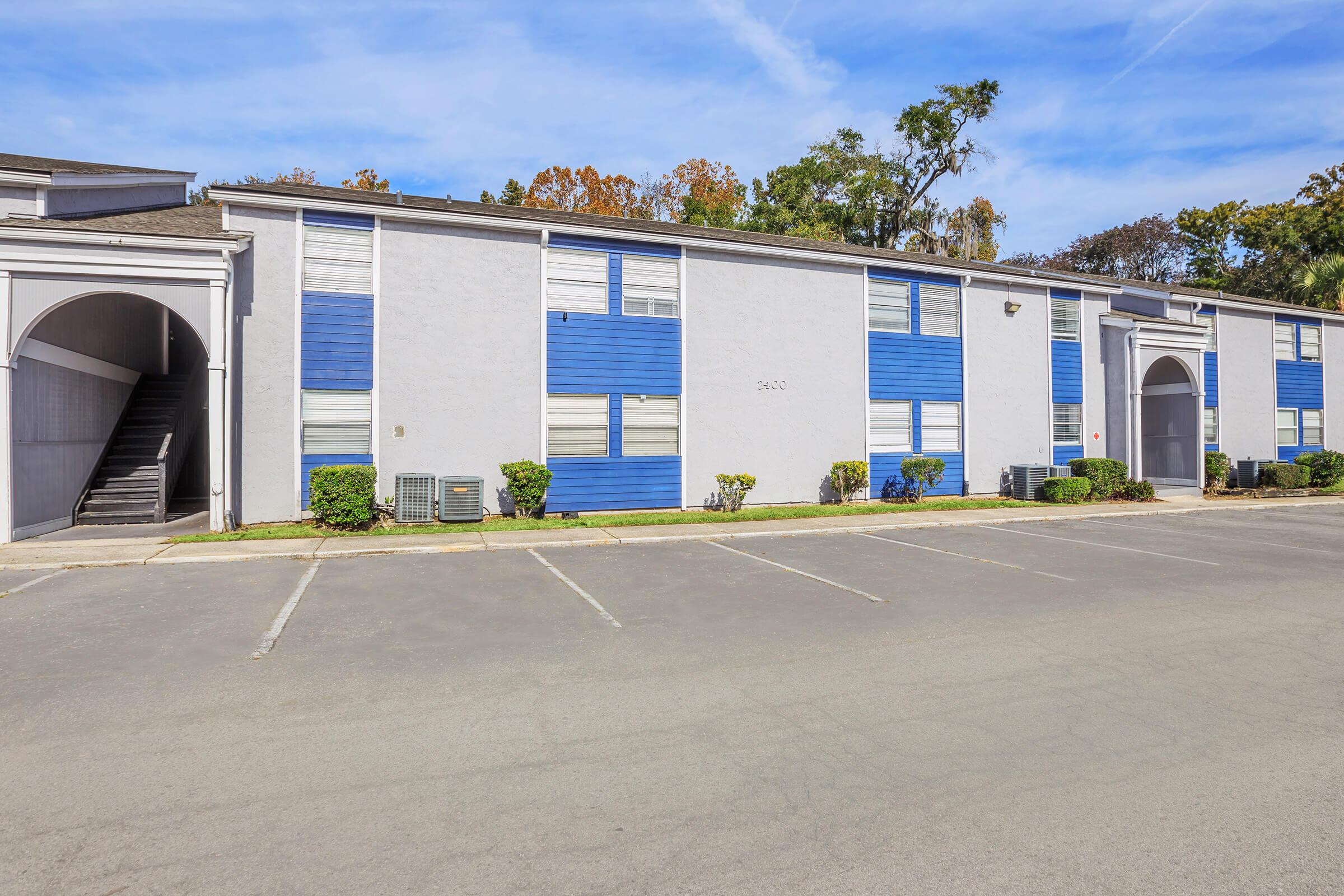 A low-rise apartment building featuring gray walls and blue accents, with a series of windows. The entrance is located under a small archway, and there are landscaped areas with shrubs in front. A parking lot with empty spaces is visible in the foreground, along with a staircase on the left side.