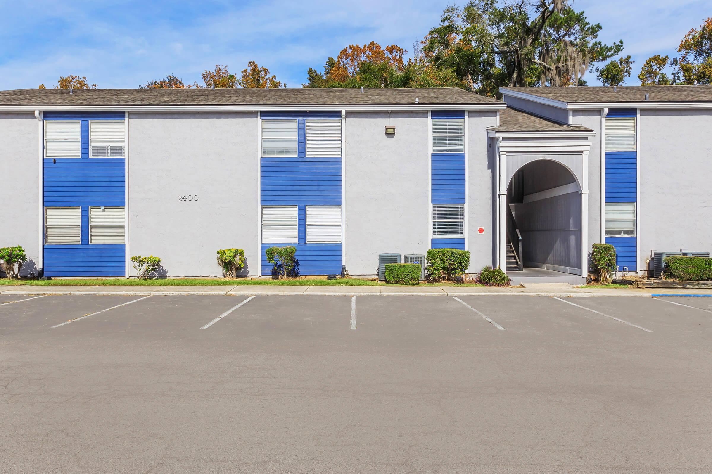 Apartment complex exterior featuring a two-story building with blue and gray siding. There are several windows, a covered entrance with an archway, and a landscaped area with small bushes. The adjacent parking lot has marked spaces. Clear blue sky in the background.