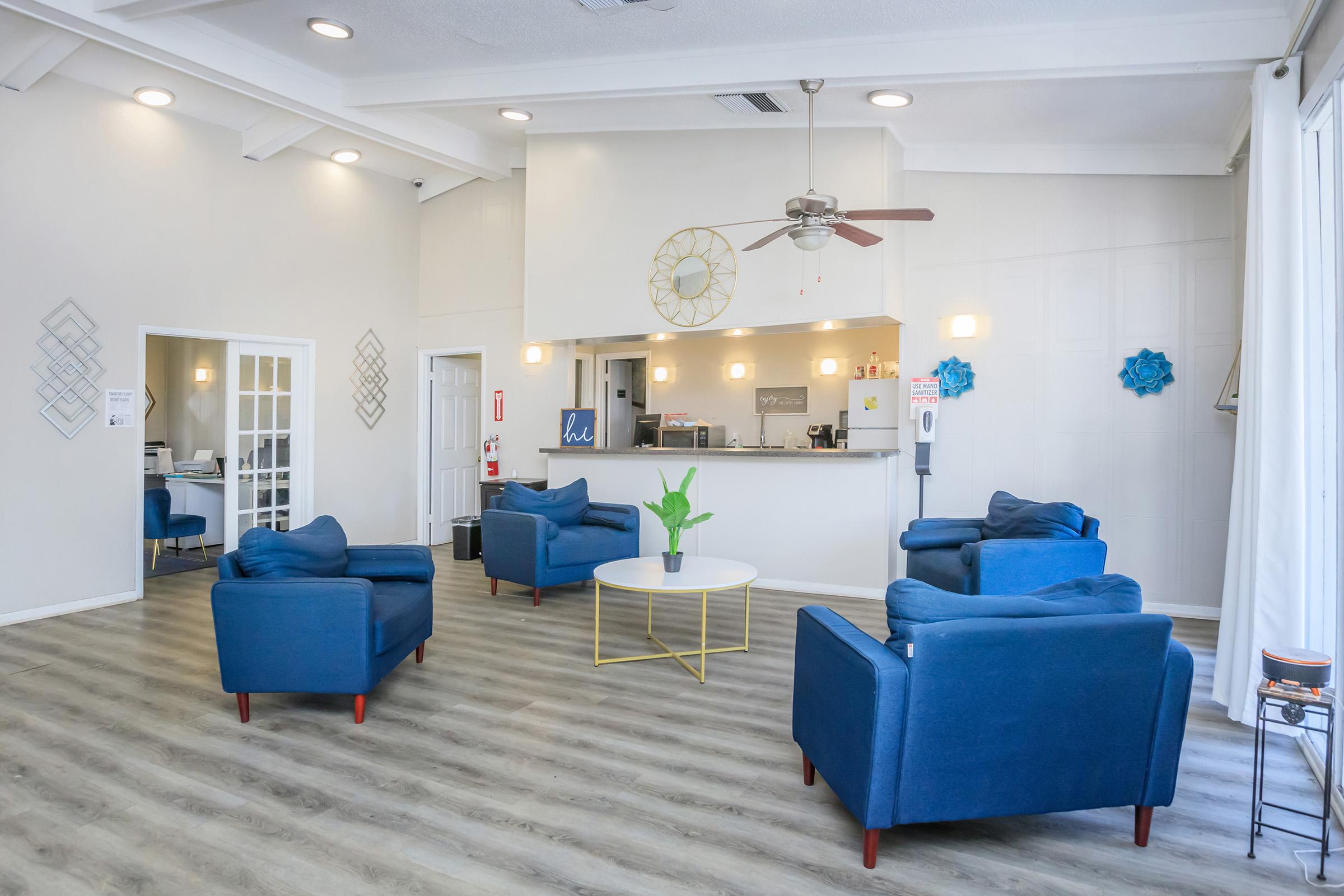 A bright, modern waiting area featuring blue armchairs arranged around a round coffee table. There is a reception counter at the back with a clock and plants. Large windows let in natural light, and decorative wall accents provide a welcoming atmosphere.