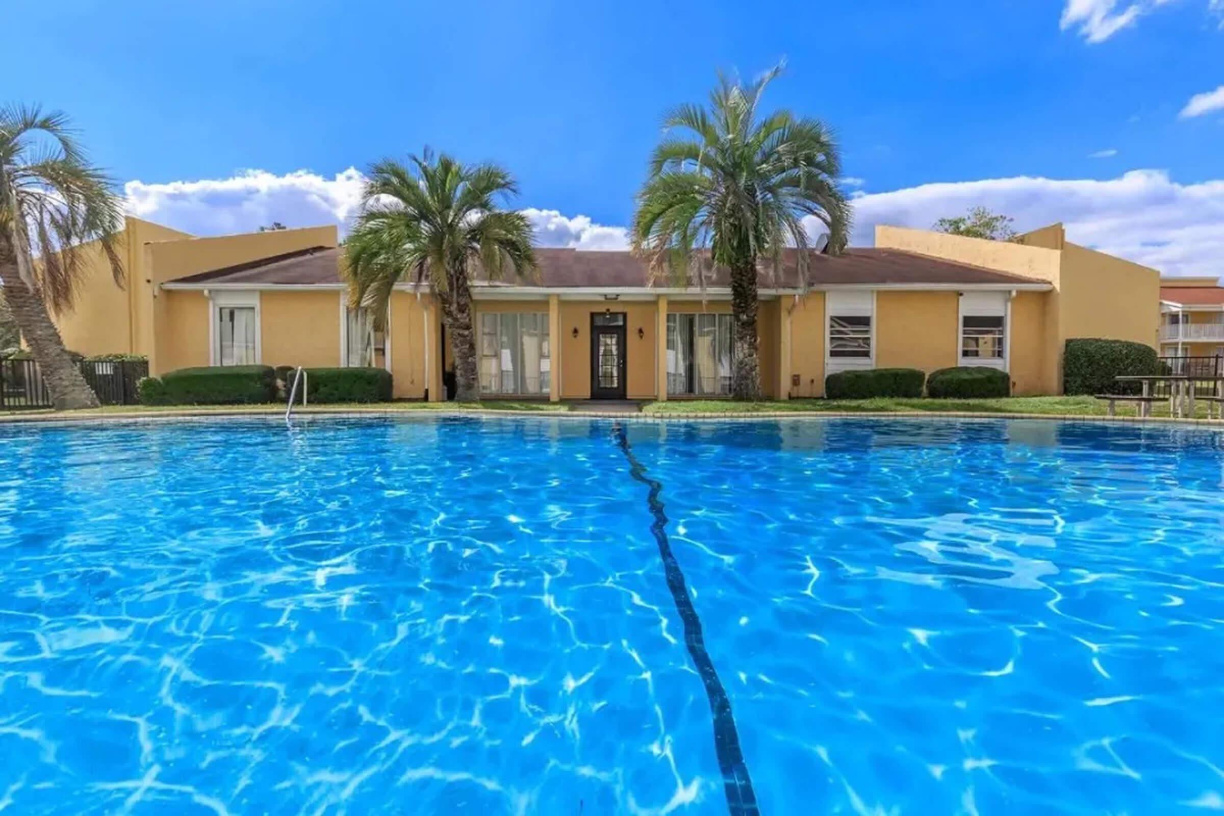 A clear blue swimming pool in the foreground with a well-maintained property featuring palm trees in the background. The building has large windows and a welcoming entrance, bathed in sunlight under a partly cloudy sky. The overall scene conveys a relaxing, tropical ambiance.
