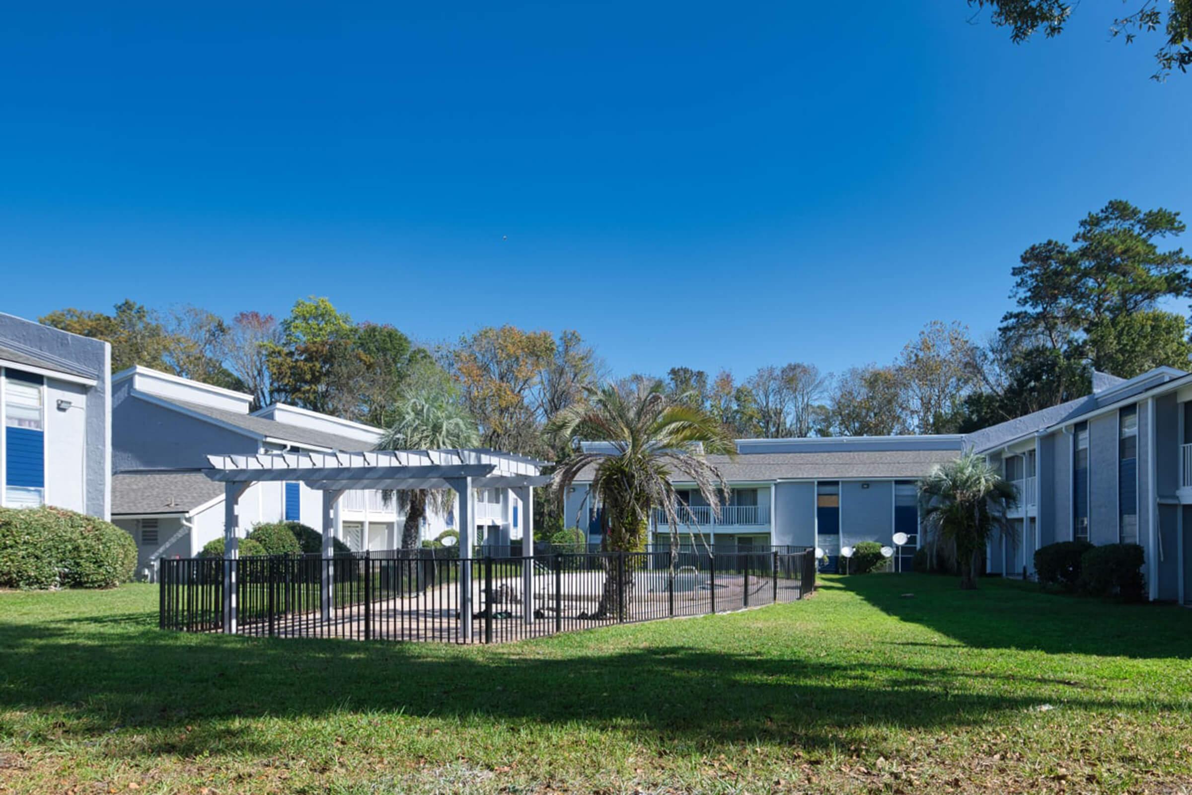 A sunny outdoor view of a residential complex featuring a fenced-in area with palm trees and grass. Surrounding buildings are two stories high, with blue skies above and trees in the background. The scene suggests a peaceful and well-maintained living environment.