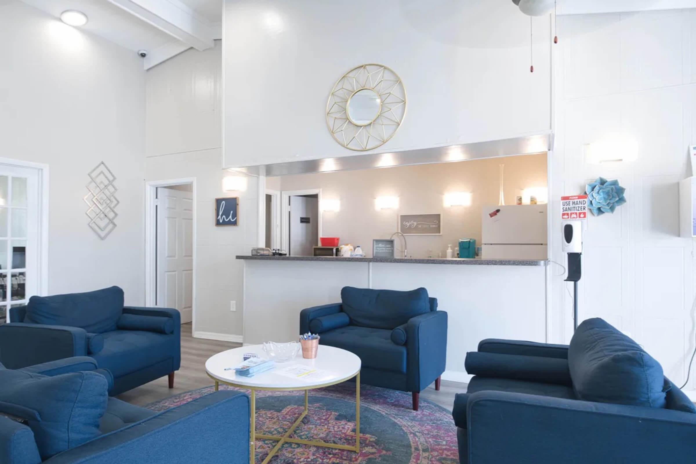 Cozy lounge area featuring four blue armchairs arranged around a round white coffee table on a patterned rug. A bar-like counter with modern decor is in the background, and a wall-mounted hand sanitizer dispenser is visible. The walls are painted white, and there's natural light from large windows.
