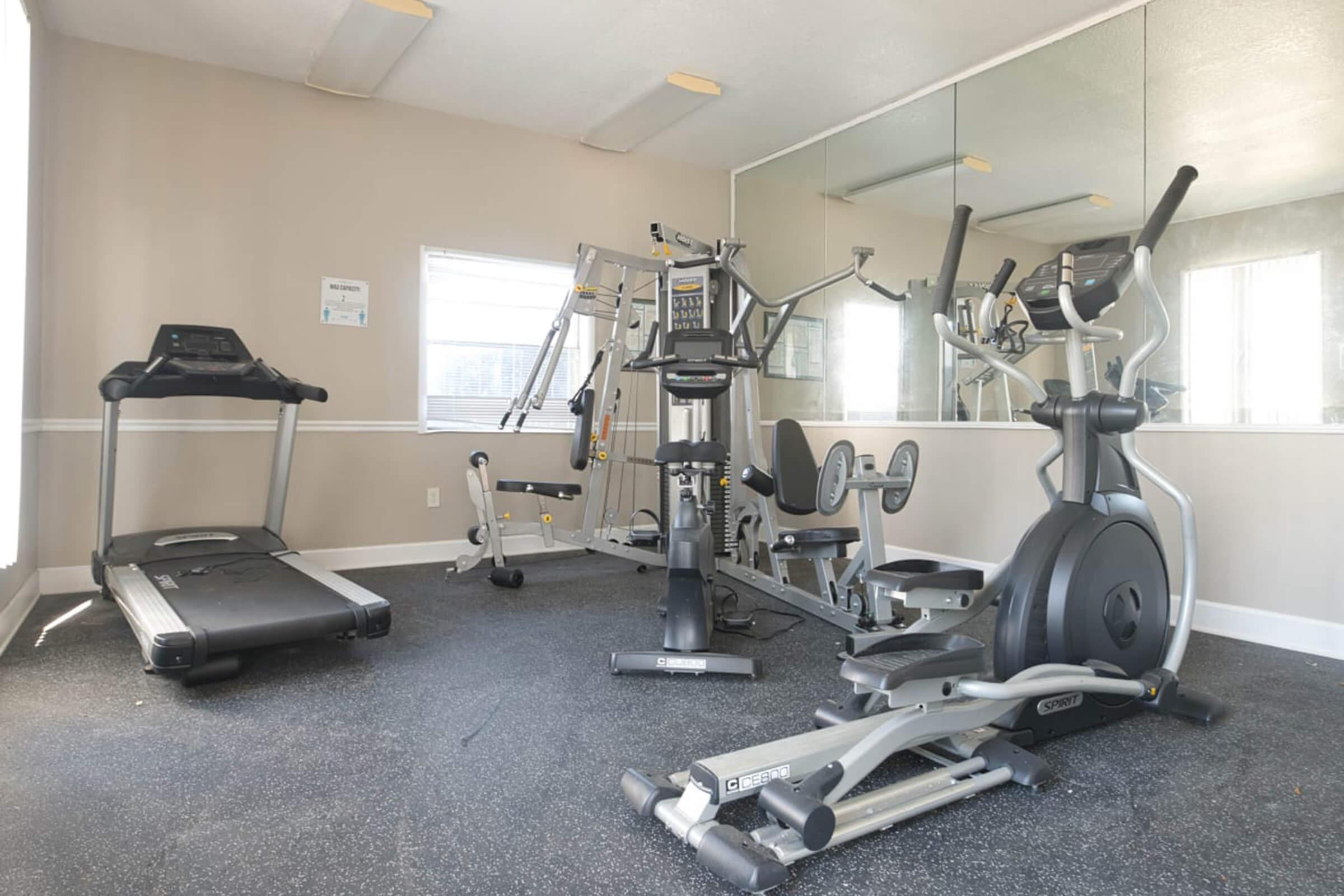 A well-lit gym interior featuring various exercise equipment: a treadmill, an elliptical machine, and a multi-station weight machine. The walls are painted in a neutral color, and there is a large mirror reflecting the equipment and windows allowing natural light in.