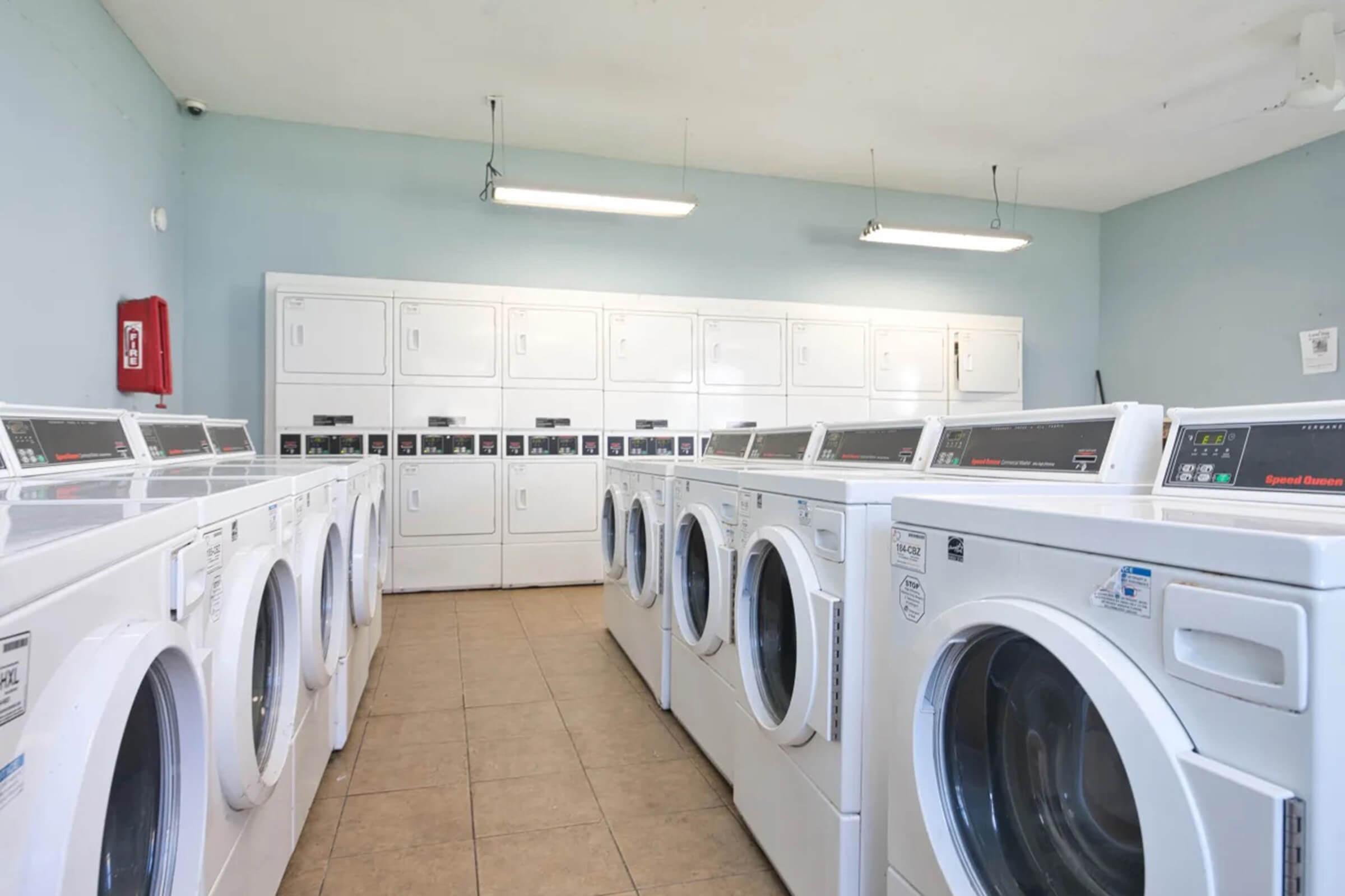 A clean and organized laundry room featuring several white washing machines and dryers aligned in rows. The walls are painted light blue, and overhead lights illuminate the space. In the background, there are cabinets for storage, and a fire extinguisher is mounted on the wall.