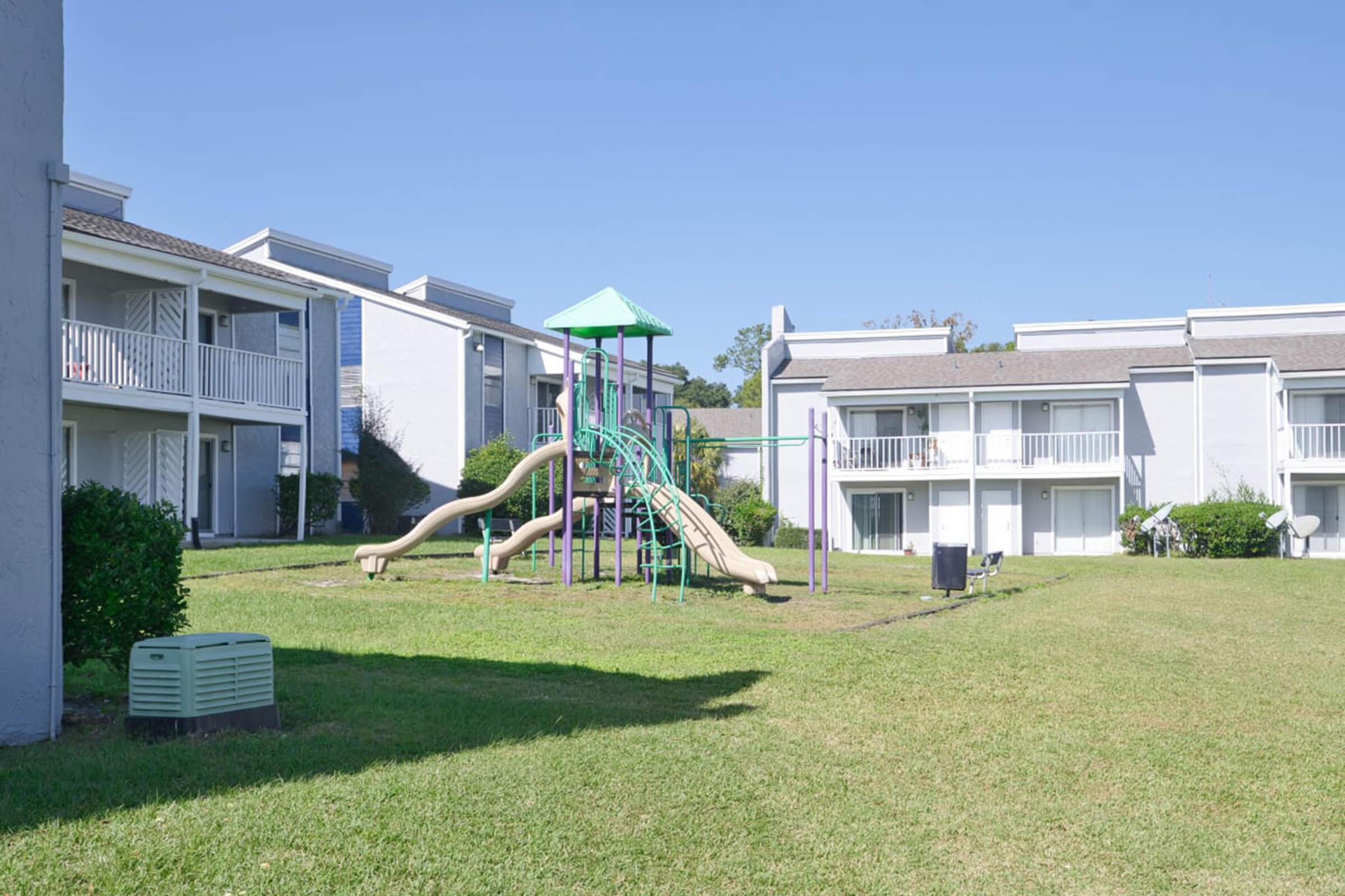 A playground featuring a slide and climbing structure, situated in a grassy area between two residential buildings. The buildings have balconies and a clear blue sky overhead, creating a sunny atmosphere.