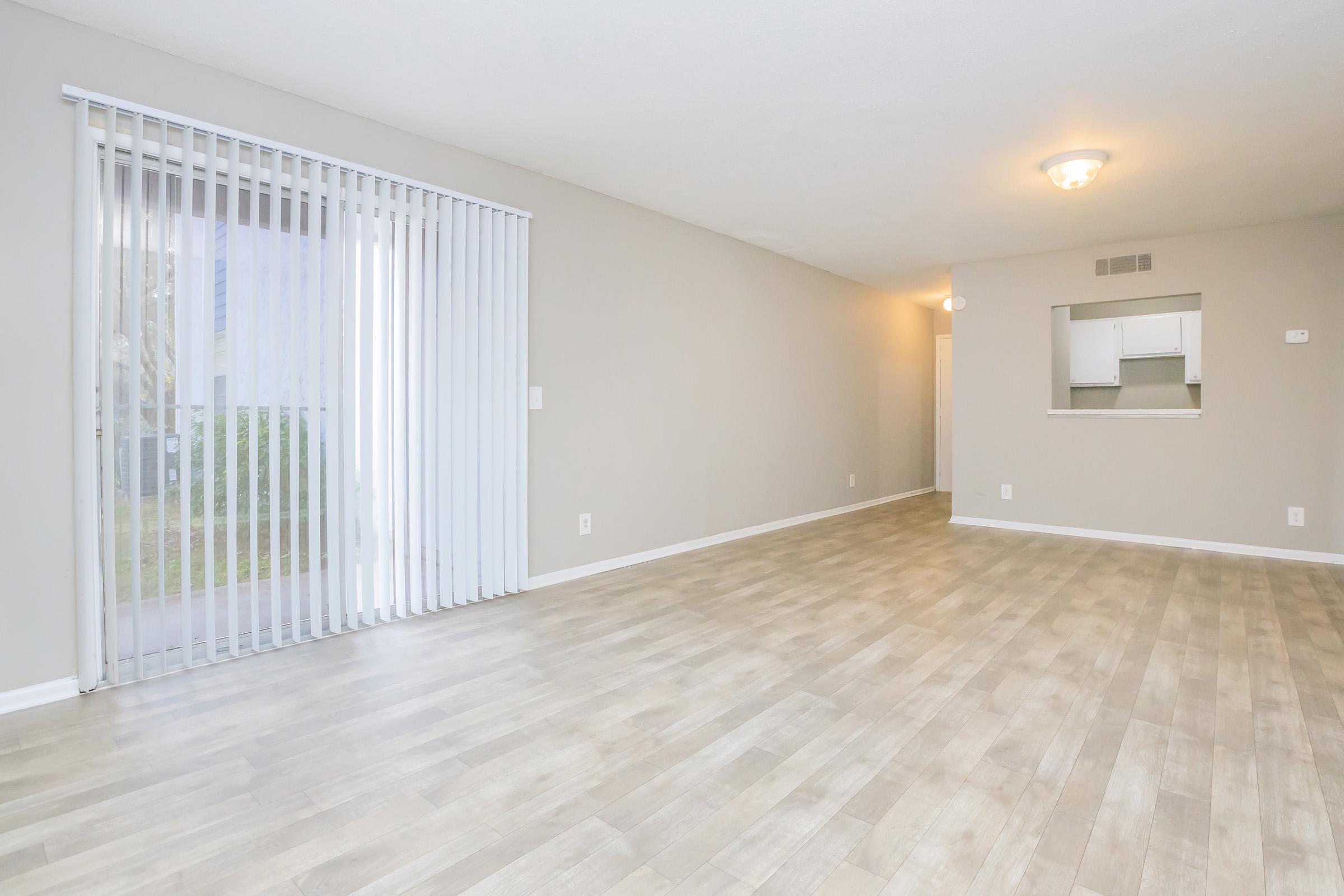 A spacious, empty living room featuring light-colored walls and wood-like flooring. Large sliding glass doors with vertical blinds provide natural light, leading to an outdoor area. A doorway in the background leads to a kitchenette, adding to the open layout of the space.