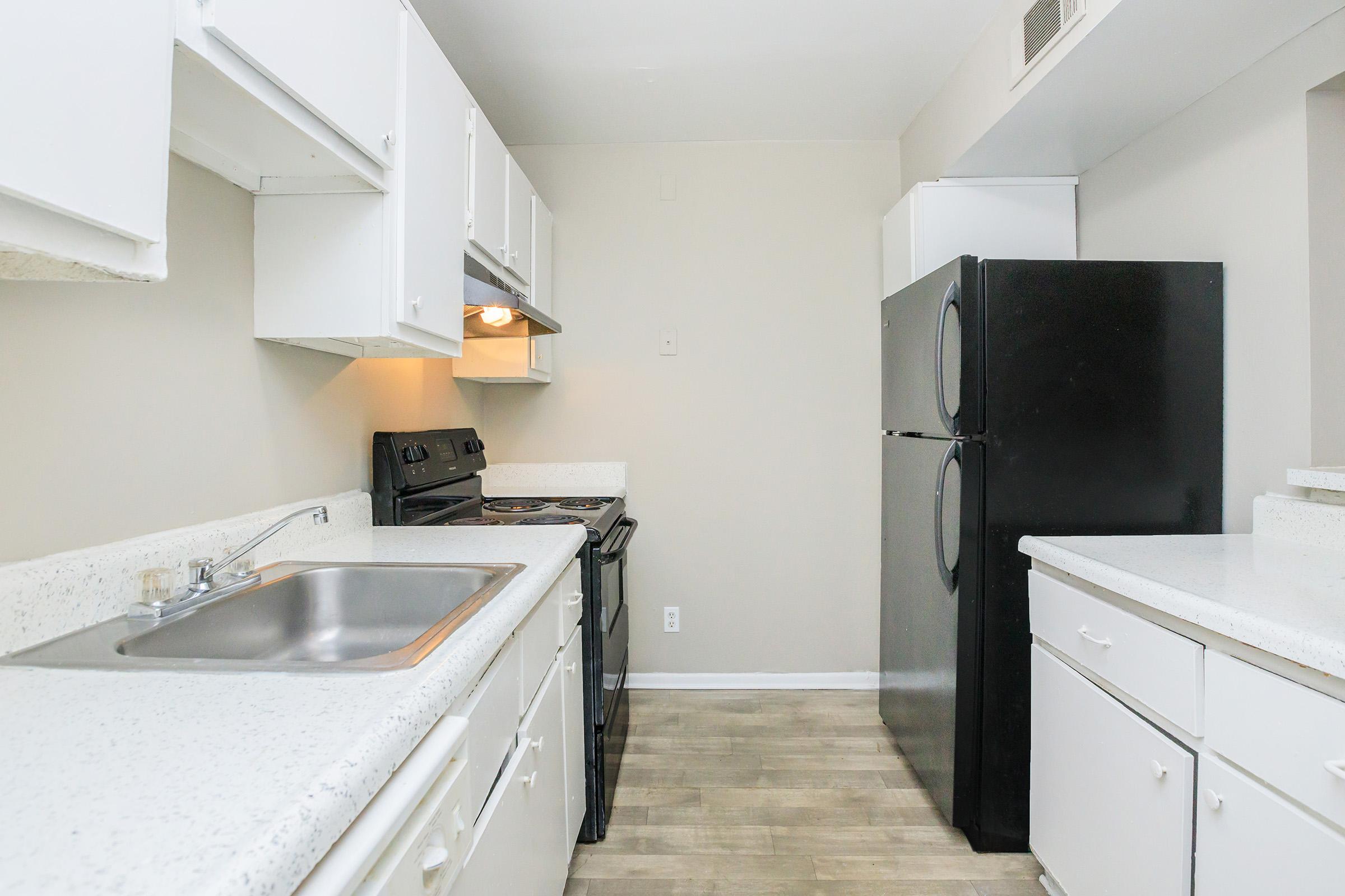 A modern kitchen featuring white cabinets, a stainless steel sink, and black appliances, including an oven and refrigerator. The countertop is a light-colored stone, and the flooring is light wood. The walls are painted in a neutral tone, creating a bright and airy atmosphere.