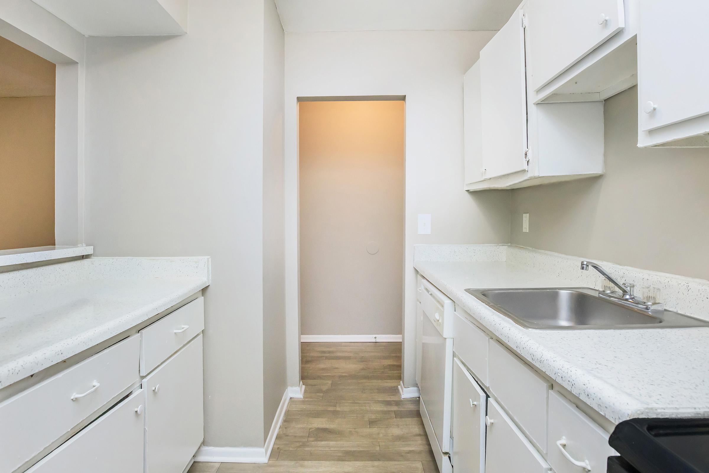 A modern kitchen featuring white cabinets, a light countertop, and a stainless steel sink. The walls are painted a neutral color, and there is a doorway leading to another room in the background. The flooring is light-colored, creating a bright and spacious feel.