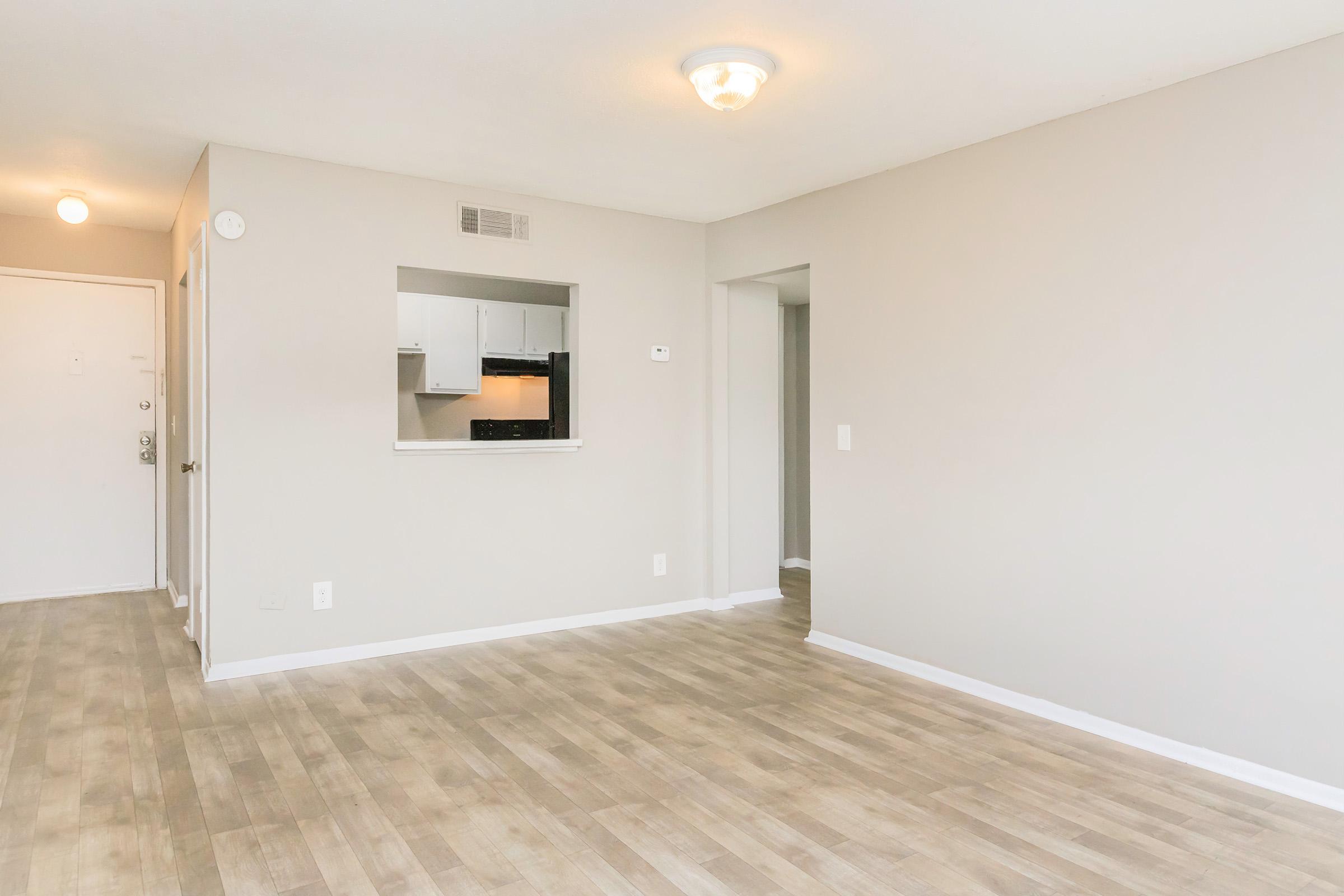 A spacious, empty living area with light-colored walls and hardwood flooring. An open kitchen area is visible through an archway, featuring a small counter. The room is well-lit with natural light and has a ceiling light fixture, providing a warm and inviting atmosphere.