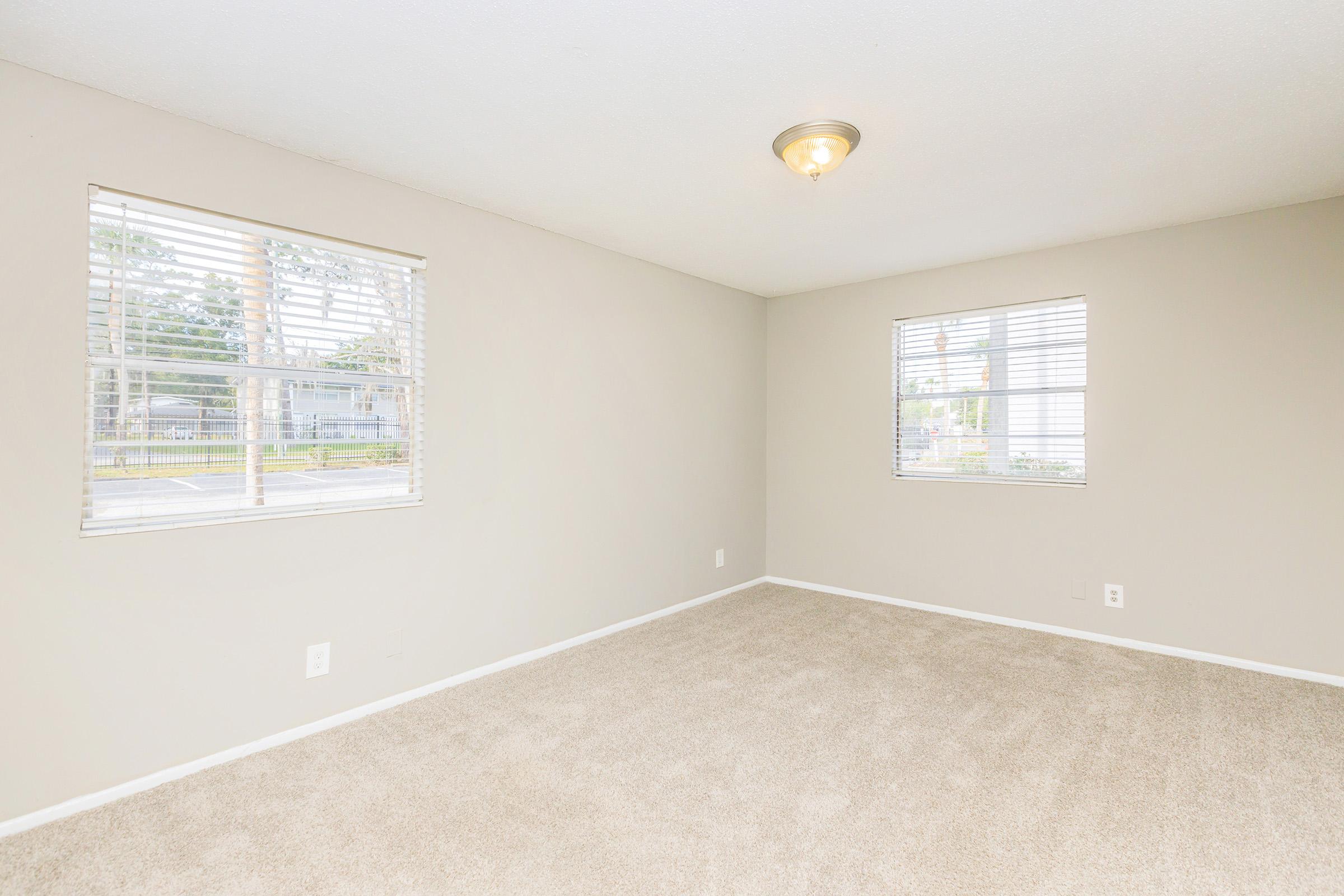 A spacious, empty room with light beige walls and soft carpet. Two windows with white blinds allow natural light to enter, illuminating the space. A ceiling light fixture adds brightness. The room appears clean and ready for use, with no furniture present.