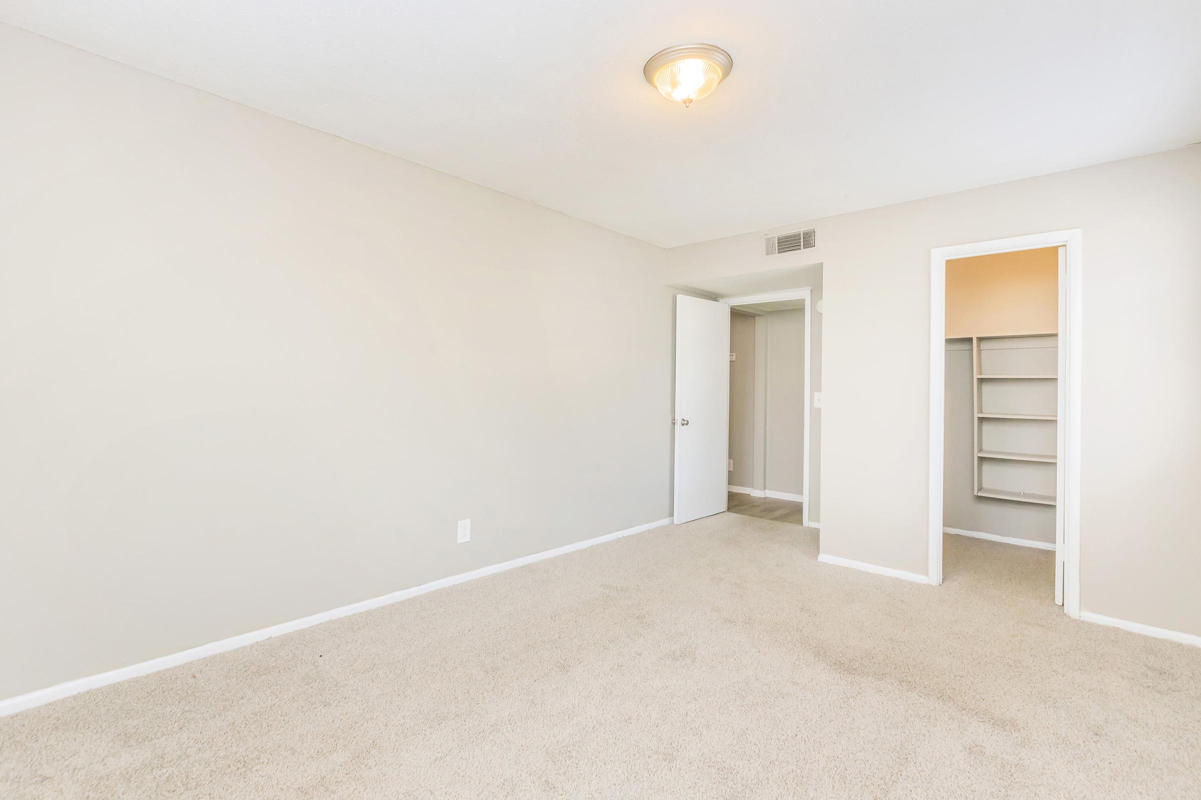 Empty room with light beige walls and light carpet. There is a ceiling light fixture, a closet with a sliding door on one side, and a small doorway leading to another area. The space is well-lit and unadorned, creating a simple and neutral atmosphere.