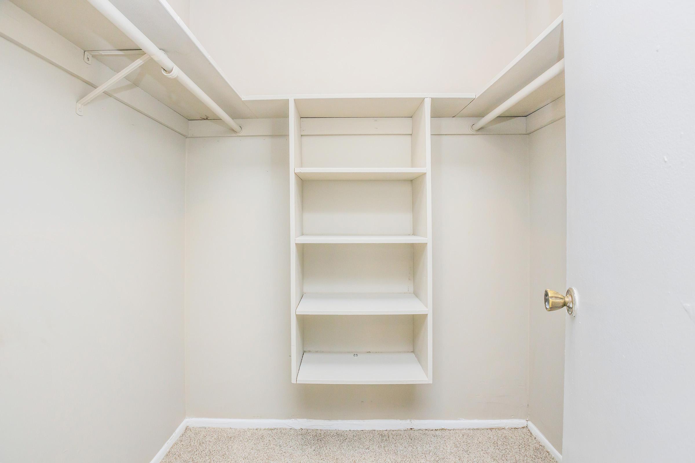 Empty closet featuring white shelves arranged on a wall, with a door handle visible on the right. The walls and ceiling are painted in a light color, and the floor is carpeted. The overall space appears clean and organized, ideal for storage or organization purposes.